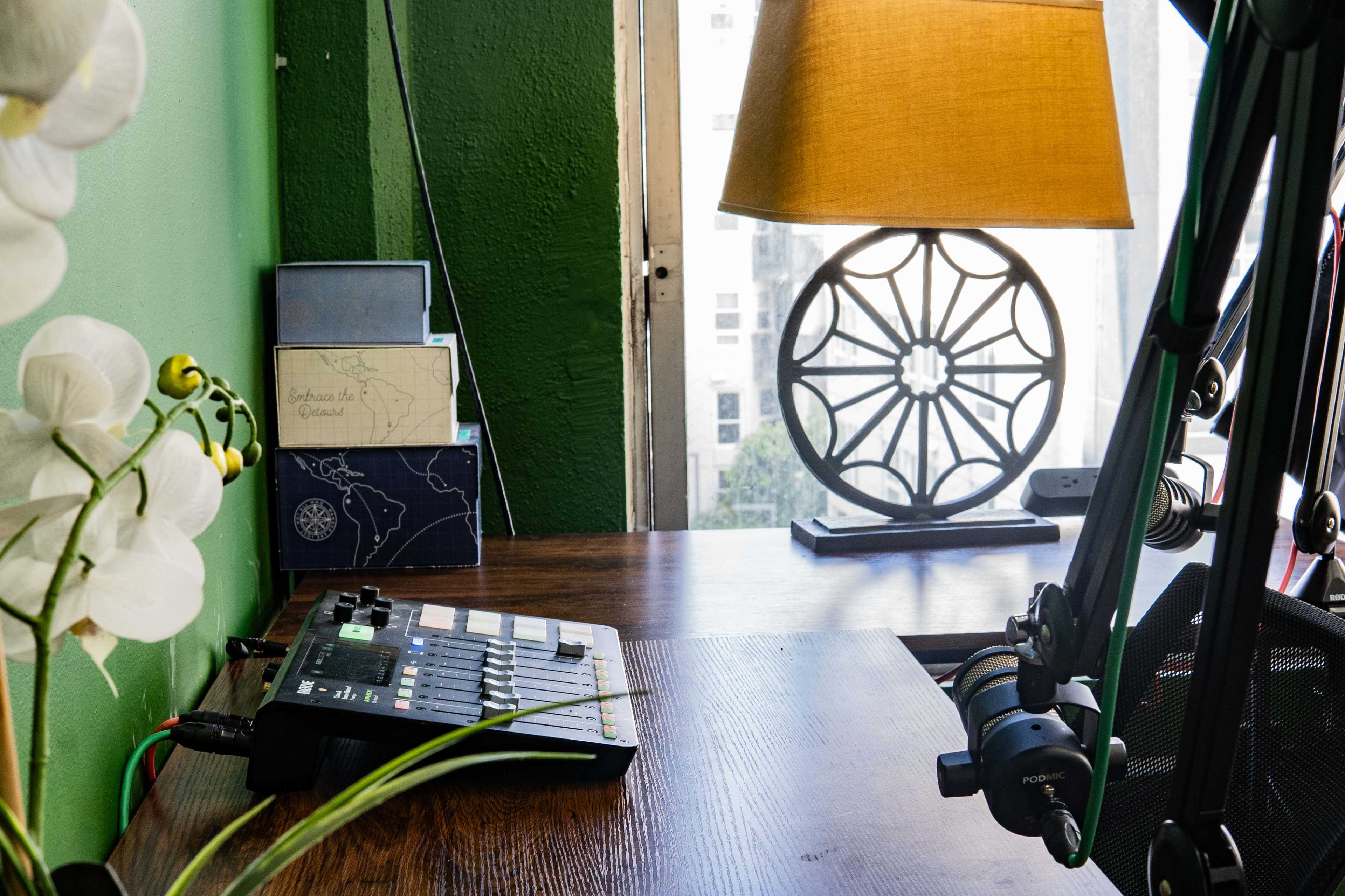 A wooden desk is set against a green wall, featuring a music production device, a lamp with a shade, and decorative elements including a plant and a window.