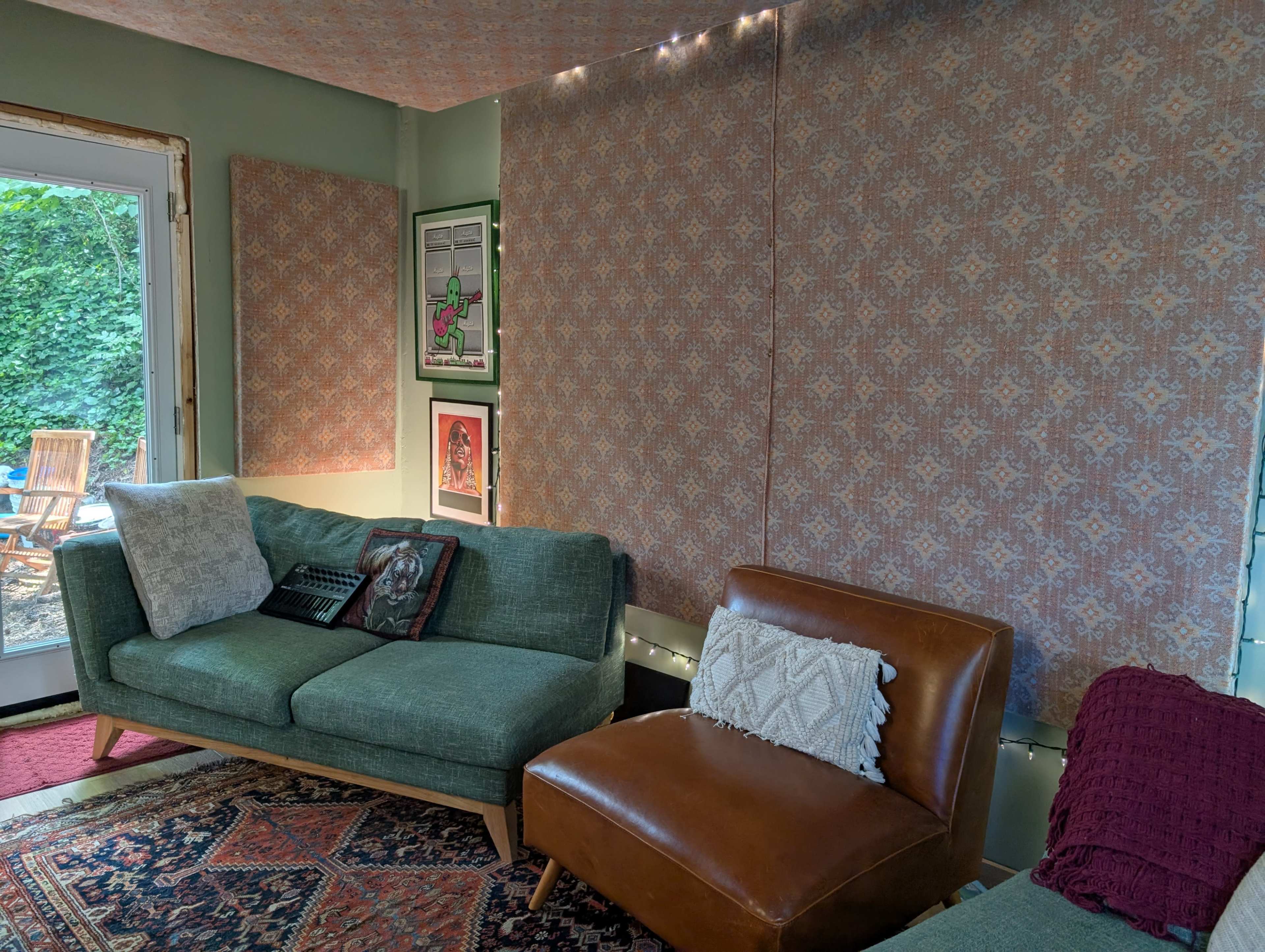 A cozy living room features green sofas, patterned wall hangings, and a doorway leading to an outdoor area with wooden chairs.