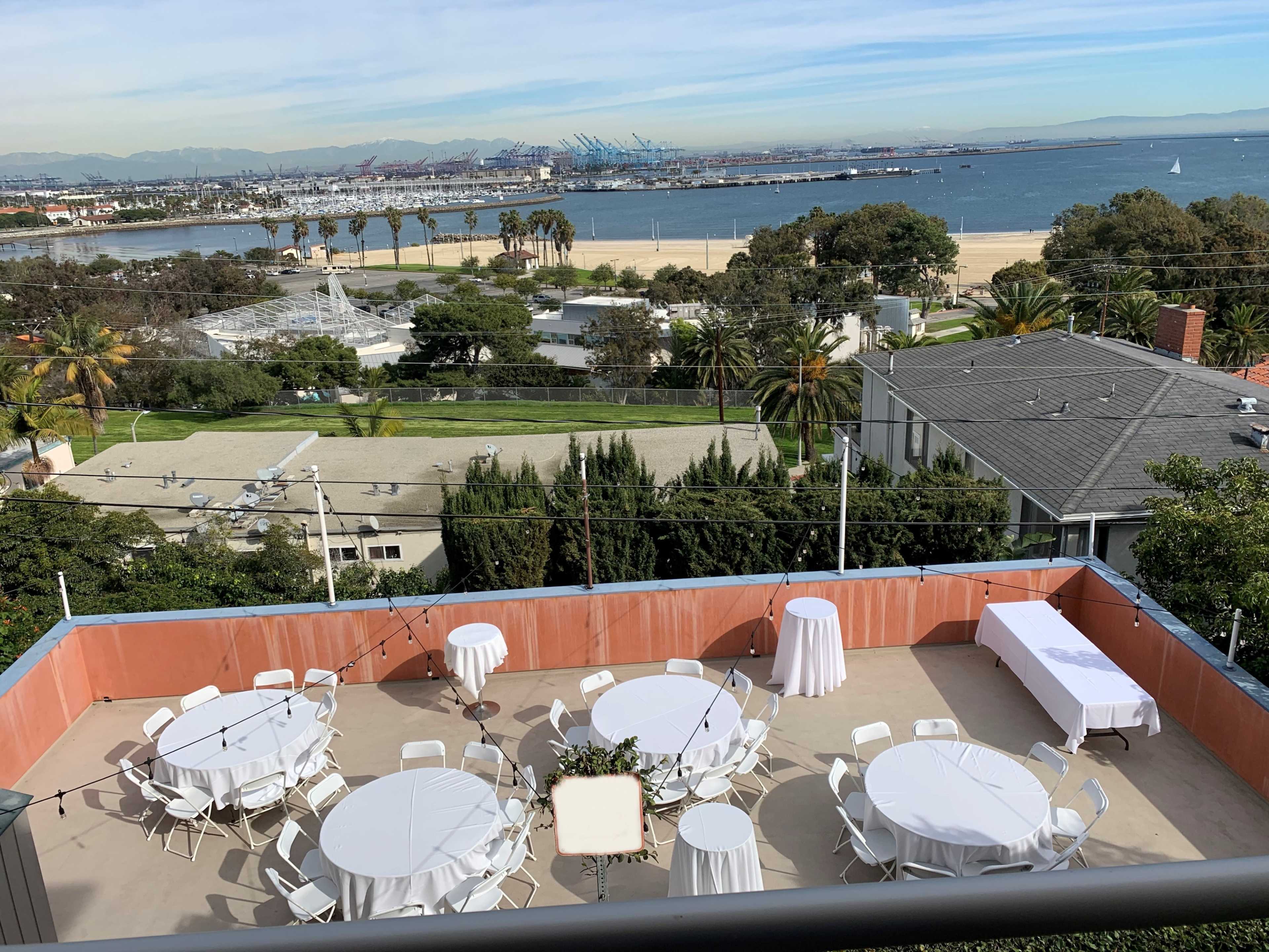 A spacious outdoor area with round tables and chairs set up for an event overlooks a waterfront and palm trees.