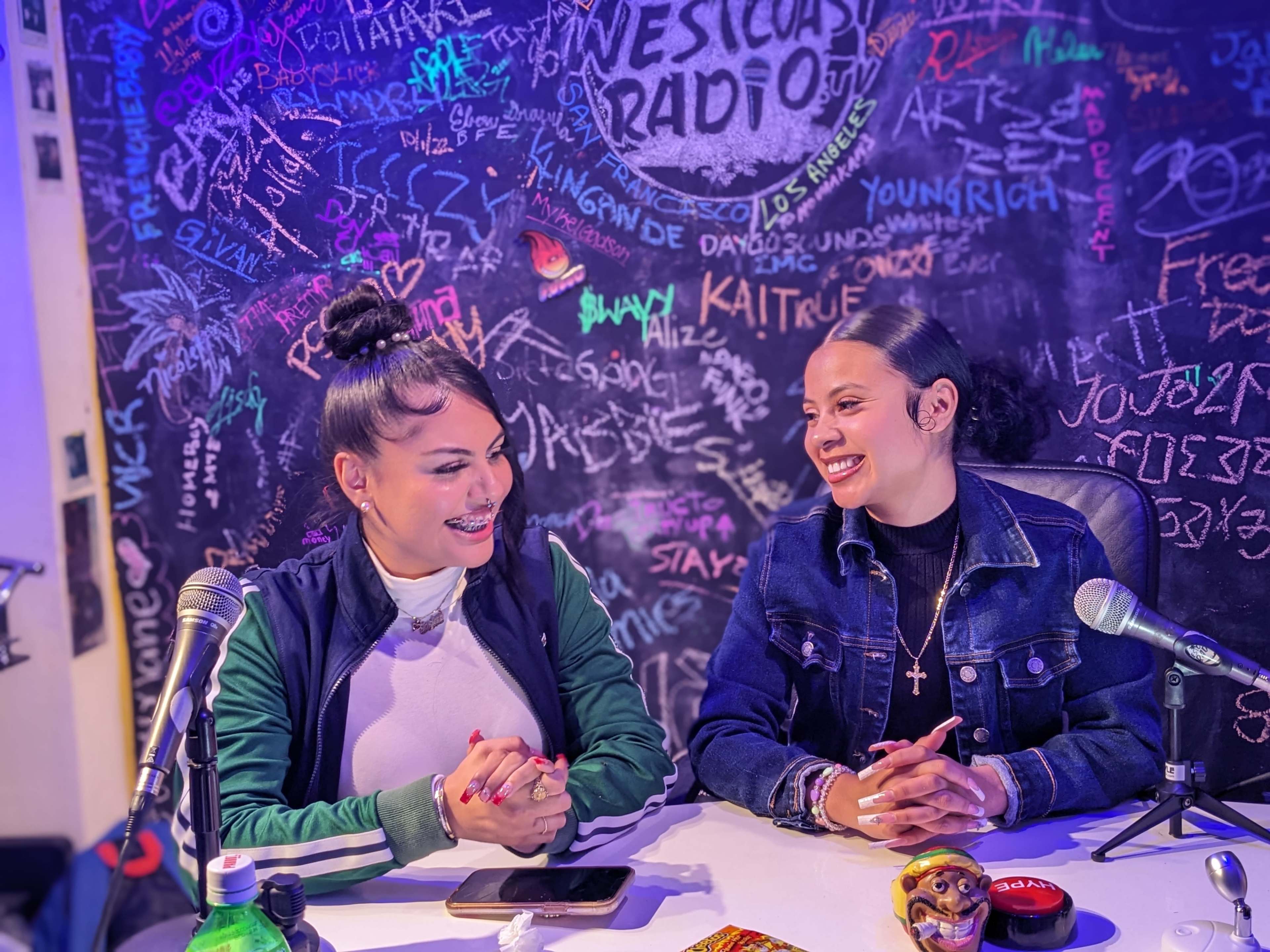 Two women sit at a table, smiling and conversing in front of a colorful graffiti-covered wall.