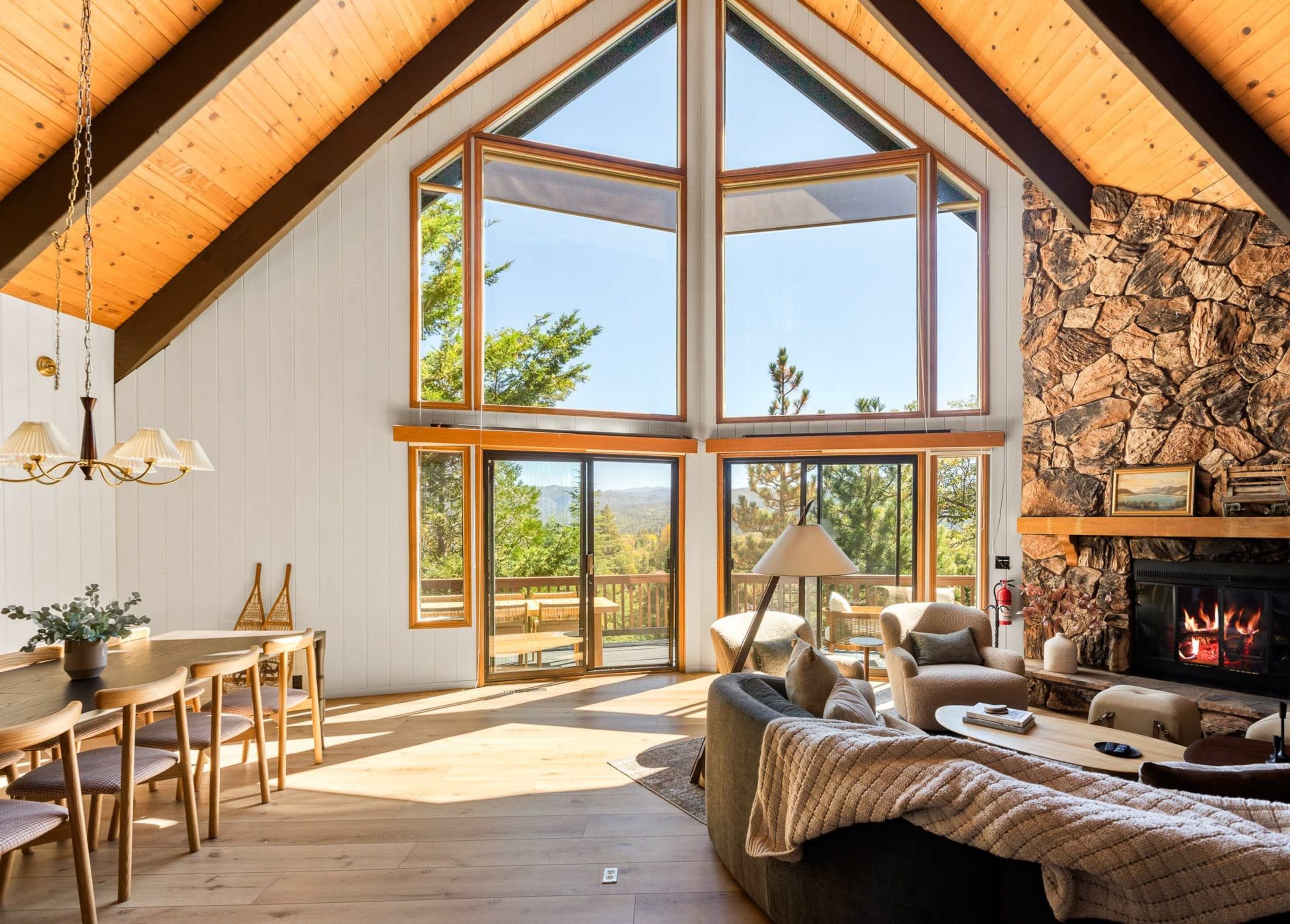 The image shows a modern living room featuring large windows, a stone fireplace, a dining area with wooden furniture, and vaulted ceilings.