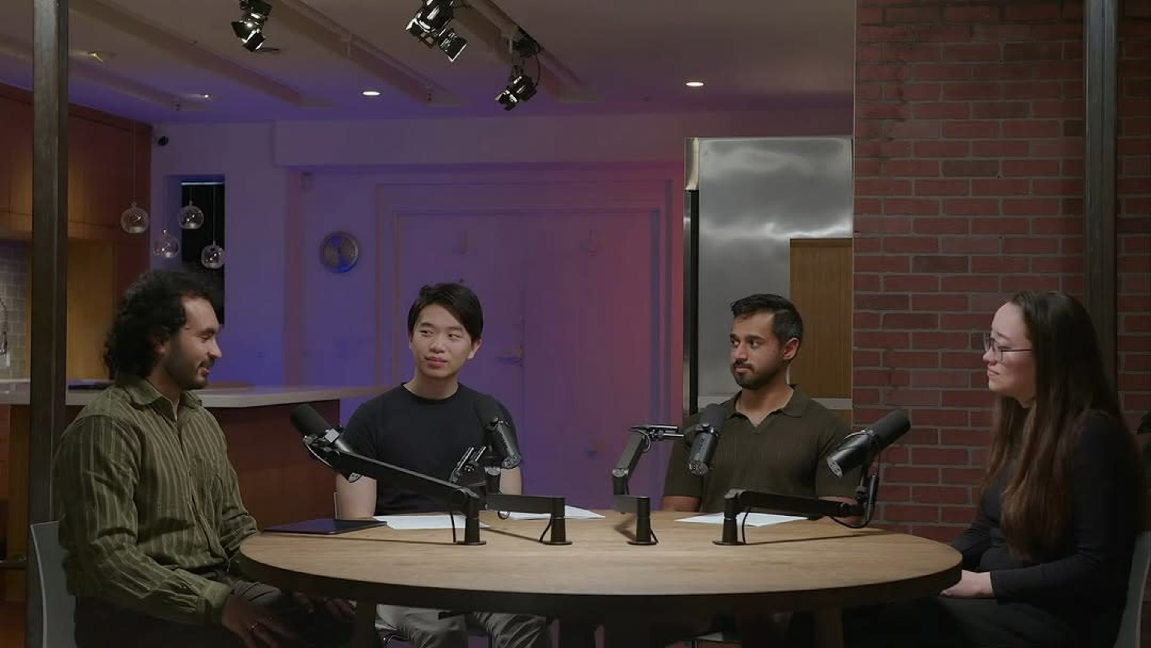 Four individuals sit at a circular table with microphones in a well-lit room featuring a modern design.