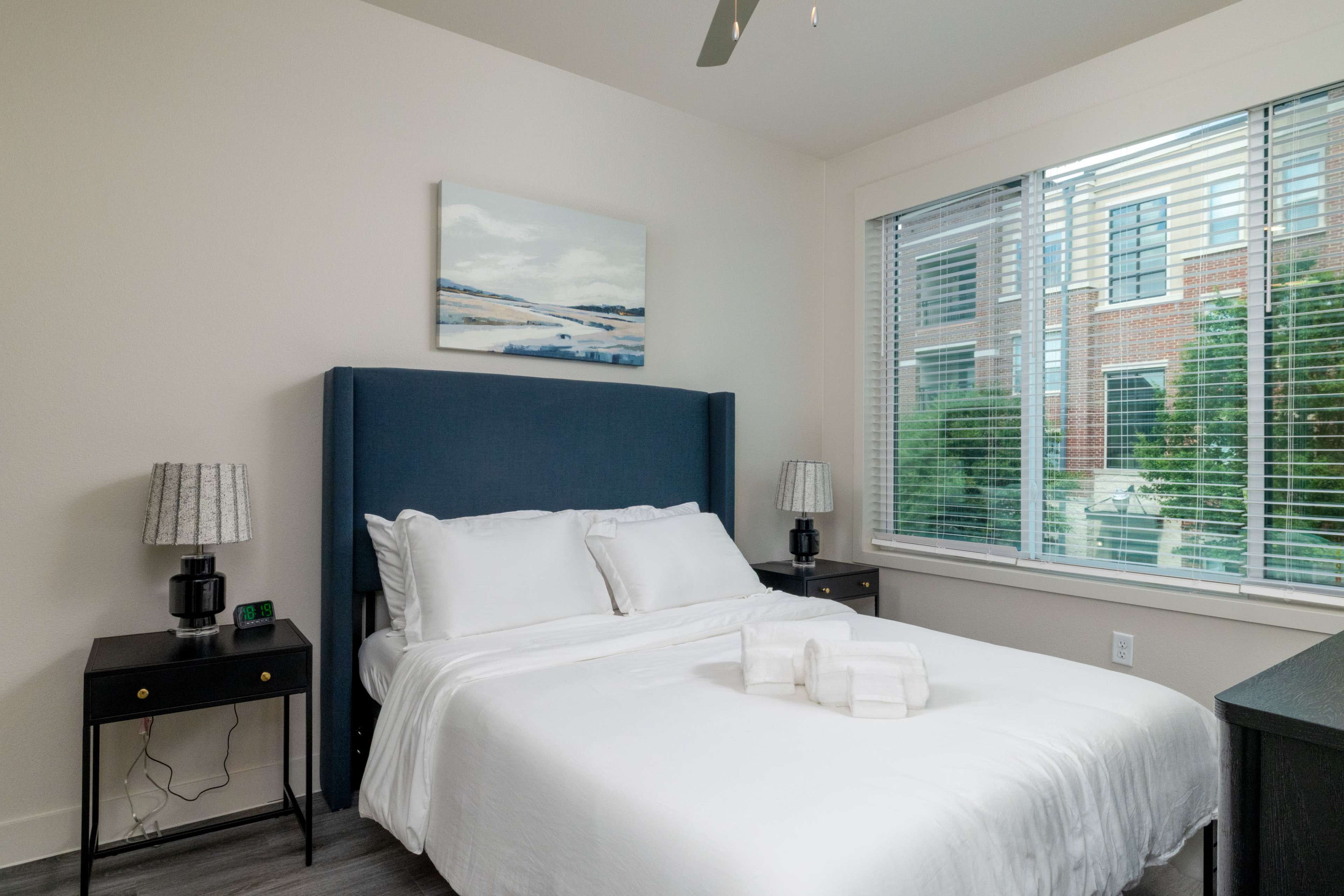 A neatly arranged bedroom features a white bed with a blue upholstered headboard, two nightstands with lamps, and large windows letting in natural light.
