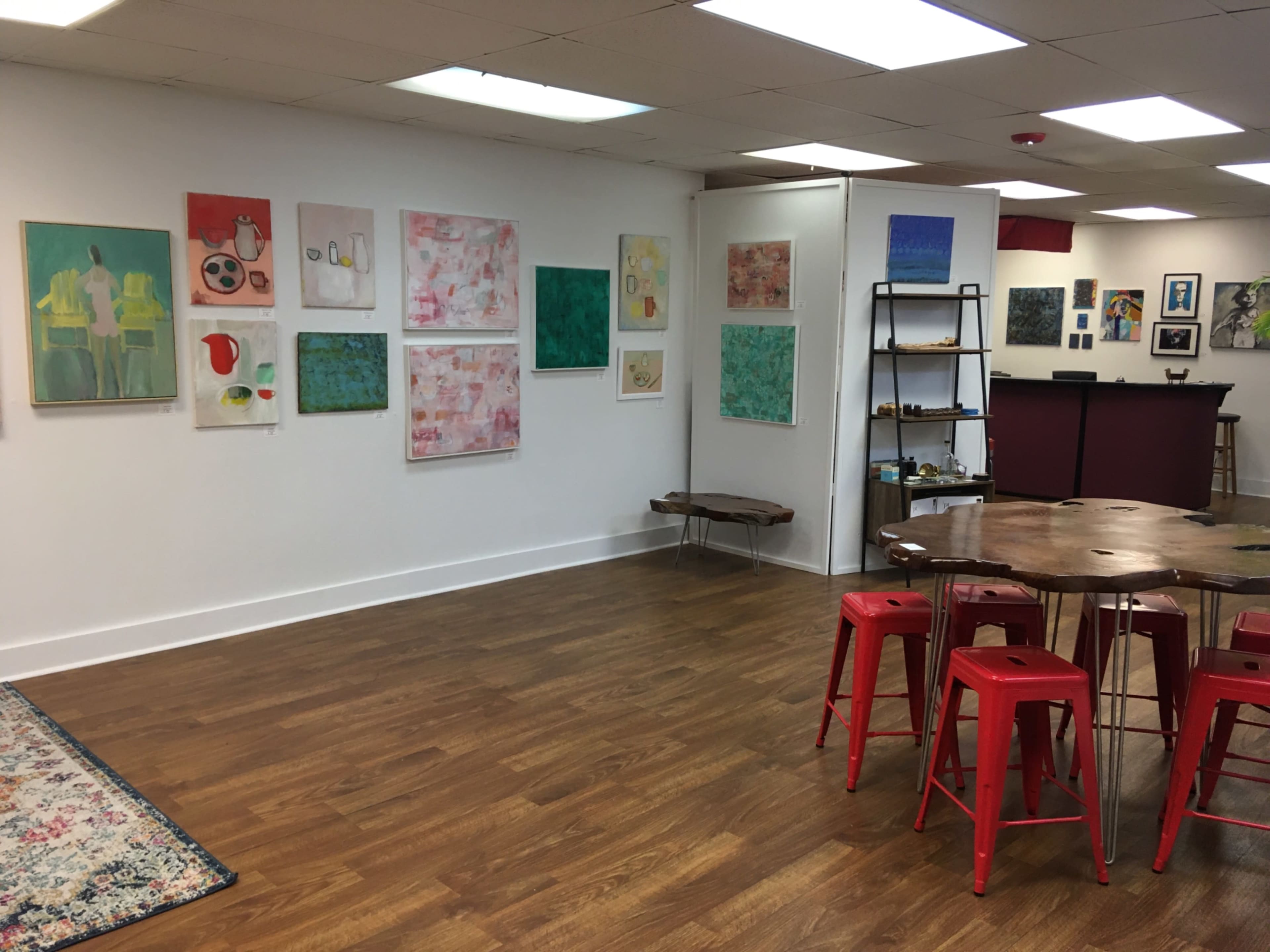 The image shows an art gallery featuring colorful paintings on the walls and a wooden table with red chairs in the center.