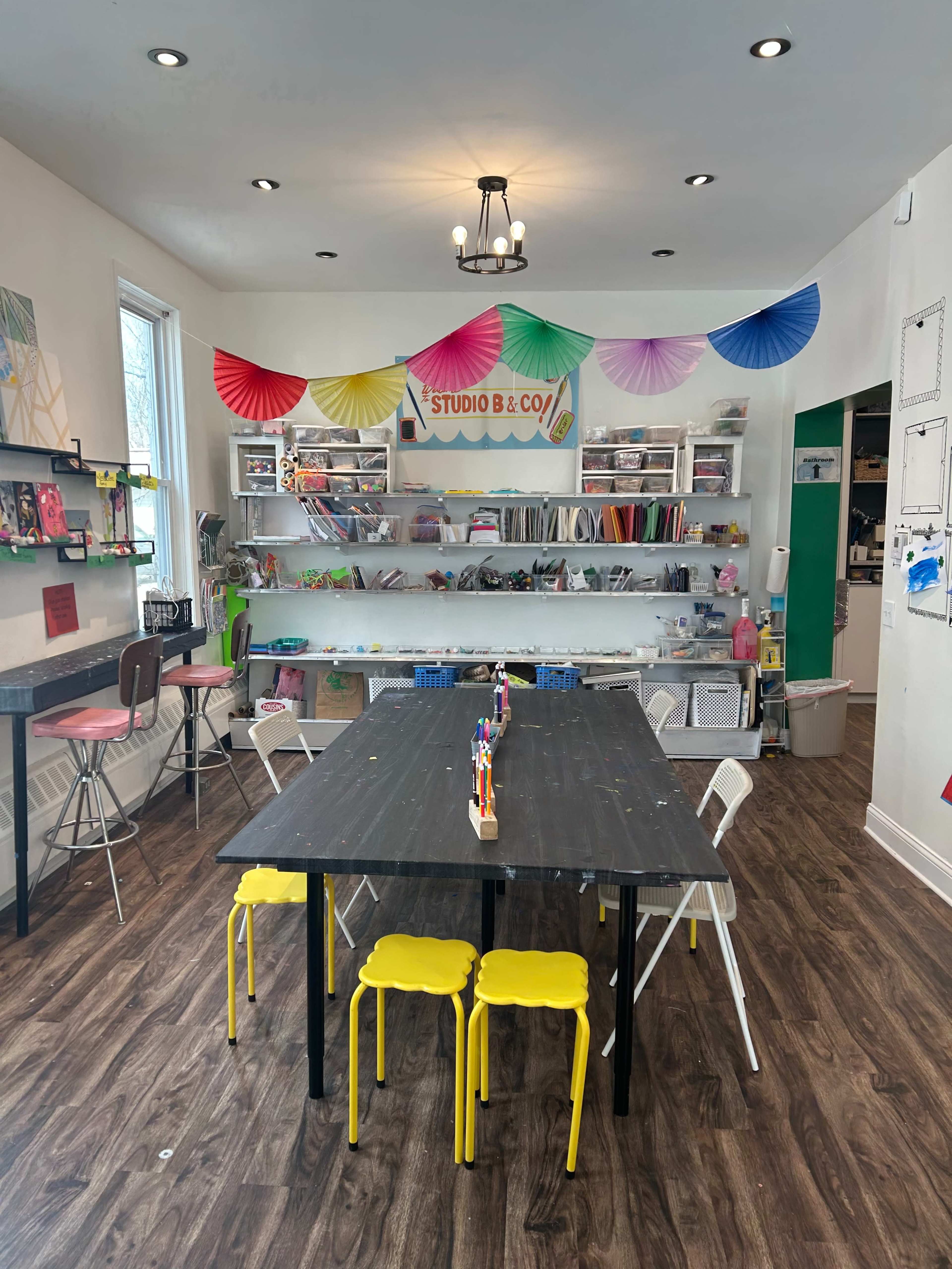 The image shows a bright and organized arts and crafts studio with a large black table in the center, surrounded by colorful chairs and shelves filled with art supplies.
