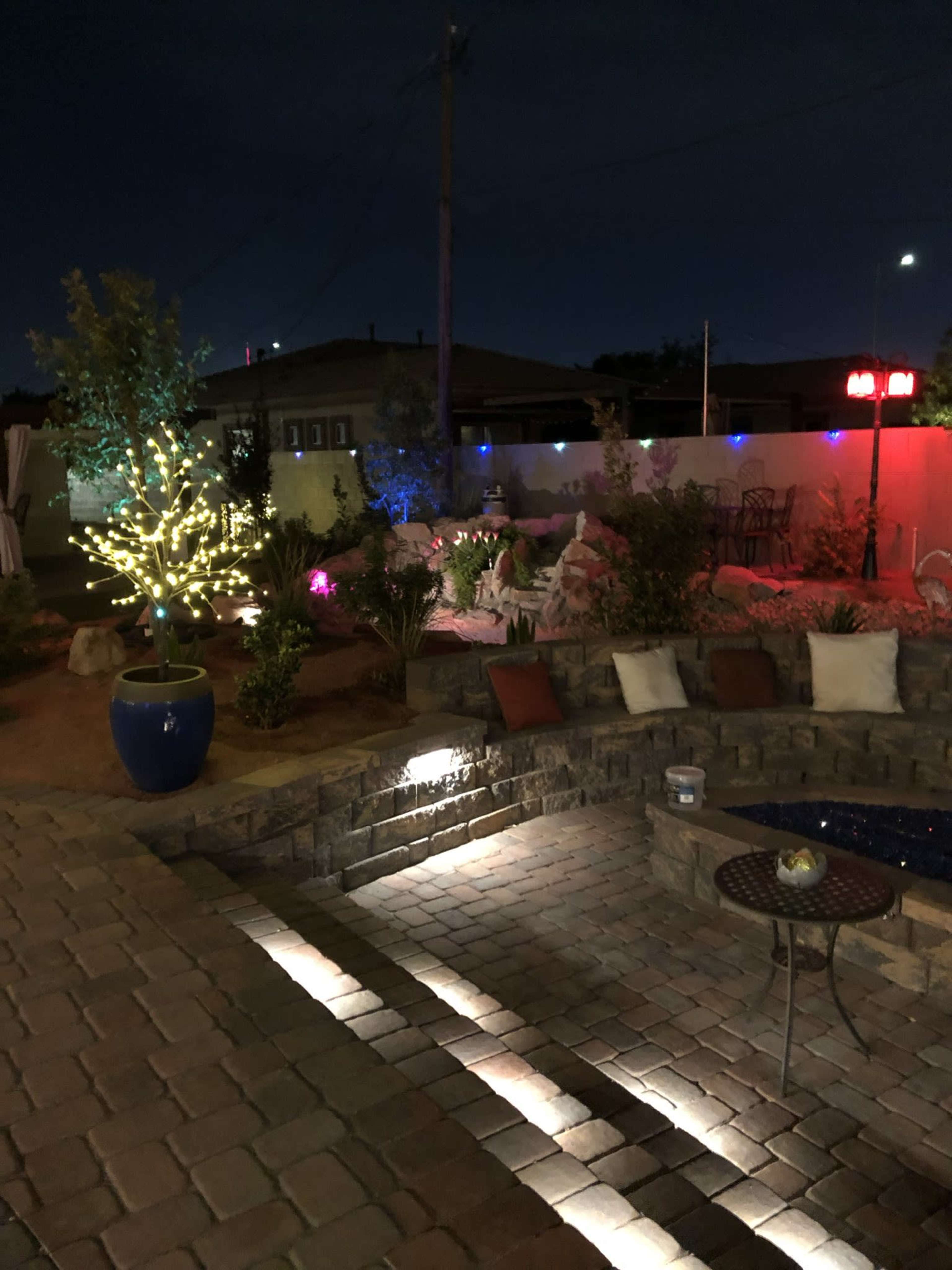 The image depicts a landscaped outdoor area at night, featuring stone pathways, decorative plants, and lights illuminating the scene.
