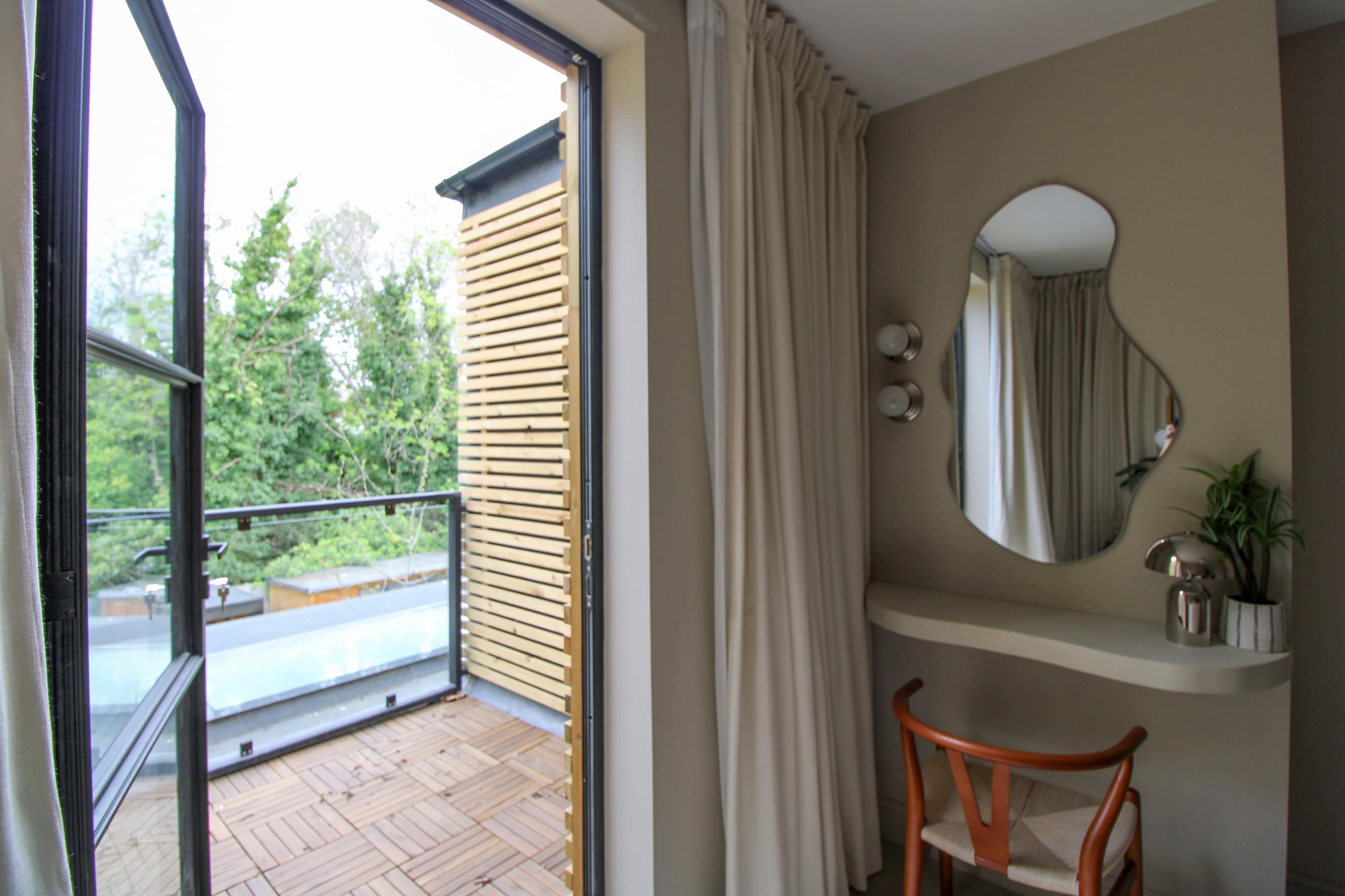 A modern room features a small desk with a mirror and a view of a balcony framed by a curtain, overlooking greenery.