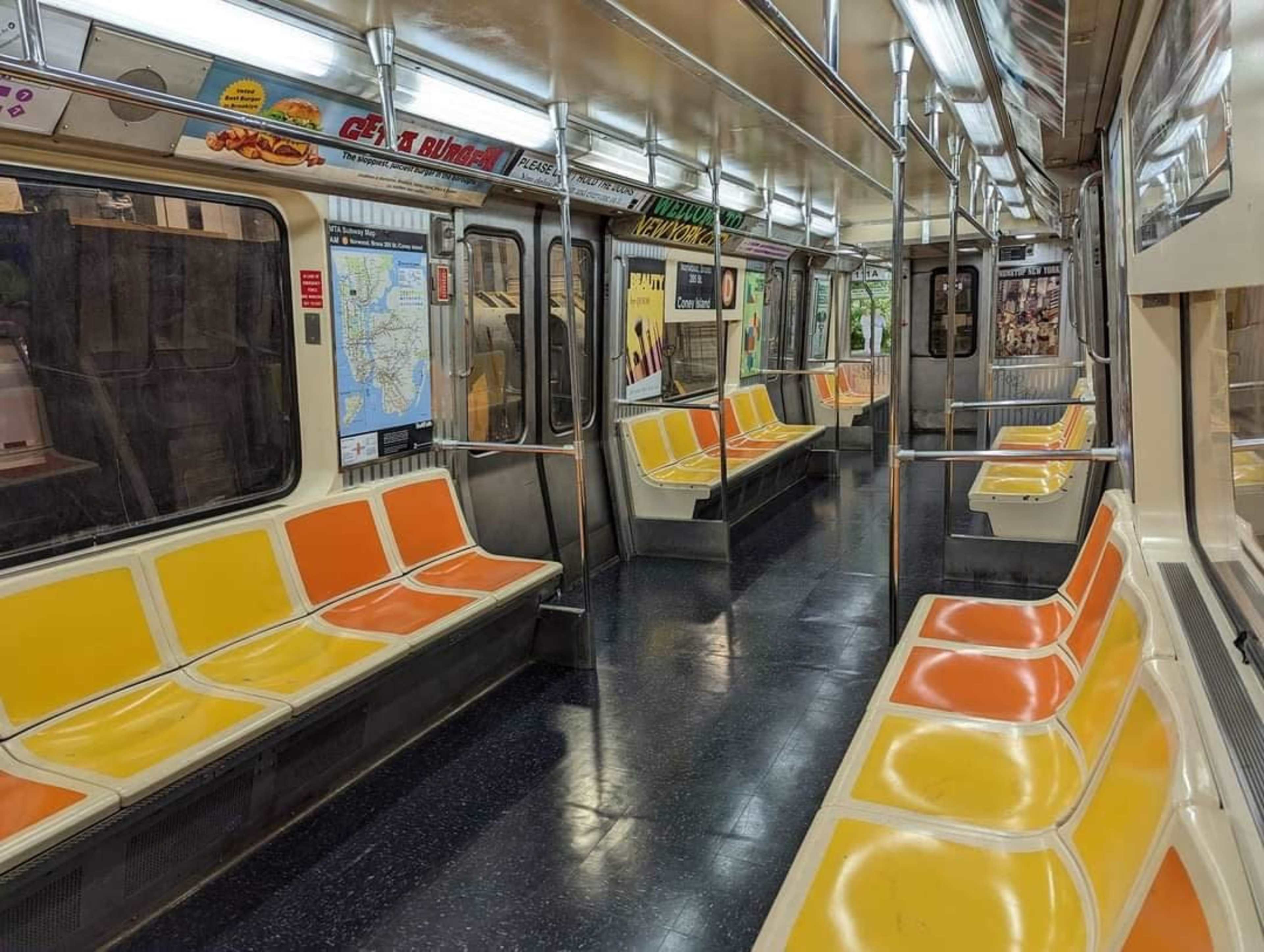 A nearly empty subway car features orange and yellow benches, with windows displaying a view of the station platform.