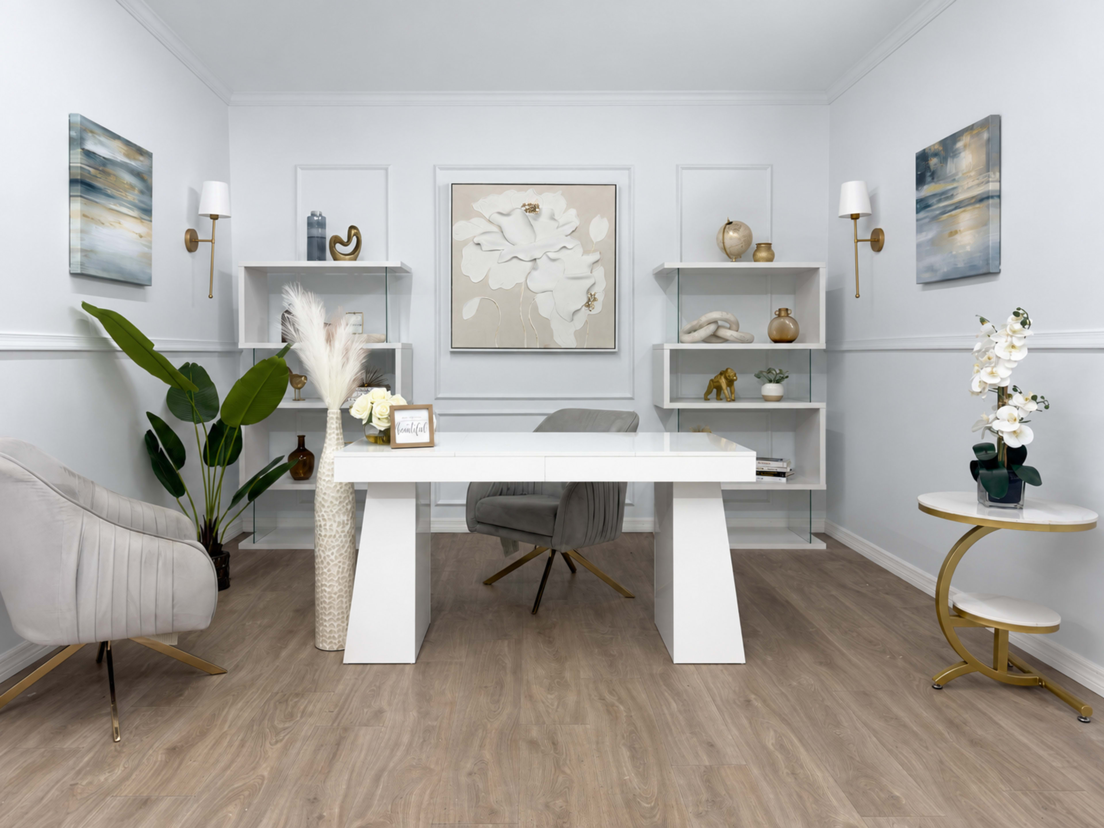 The image shows a modern office space featuring a white desk, gray chairs, shelves with decorative items, and potted plants against a light-colored wall.
