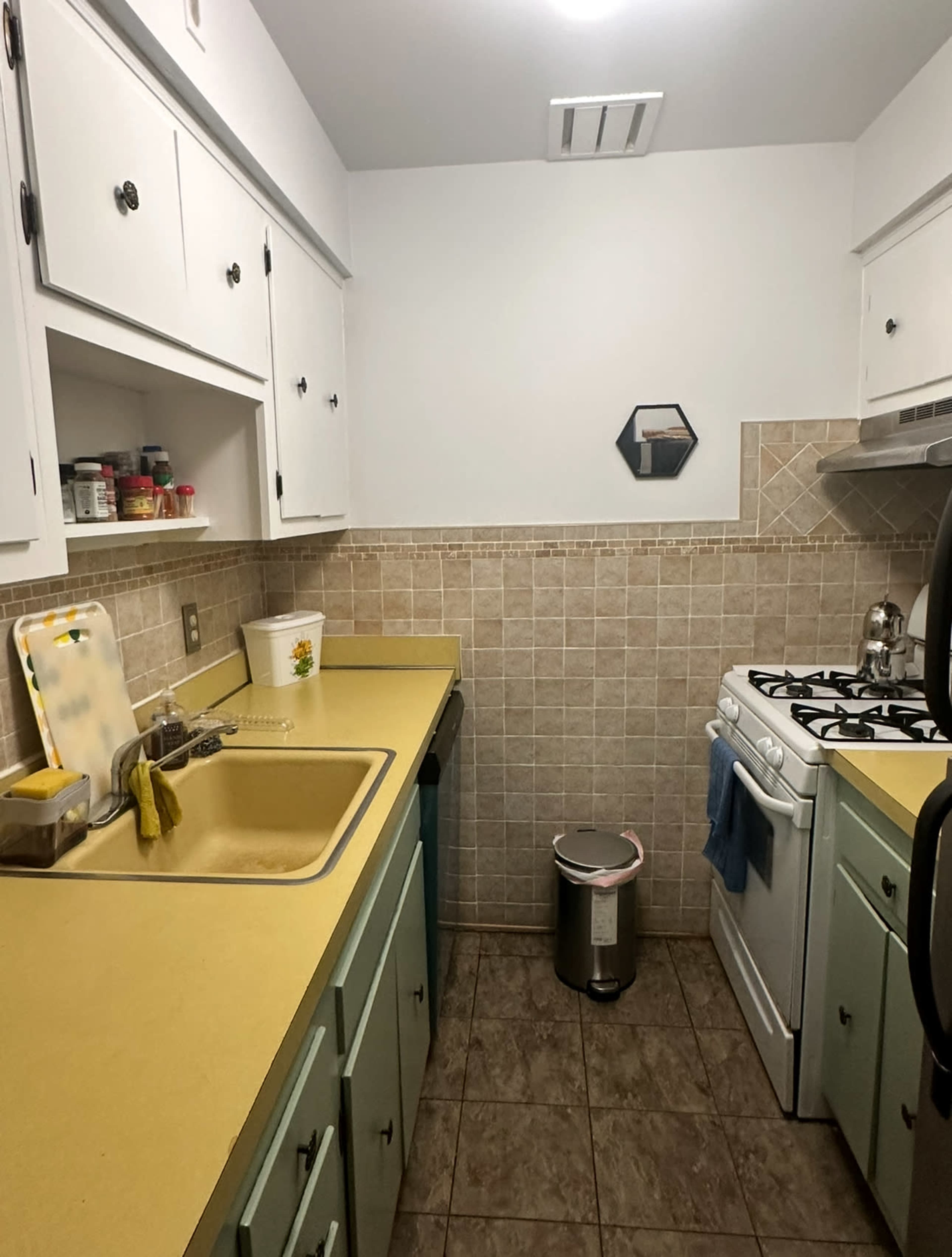 The image shows a small kitchen with a yellow countertop, tiled backsplash, and a gas stovetop next to a trash can.