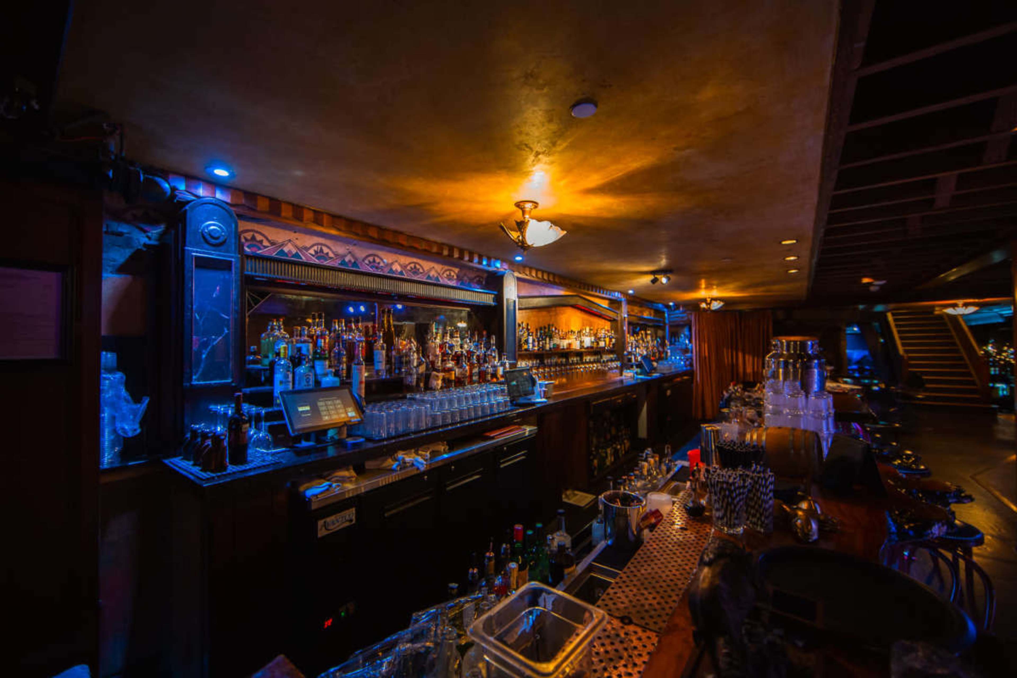 A dimly lit bar features a long counter filled with an array of bottles and glassware against a backdrop of illuminated shelves.