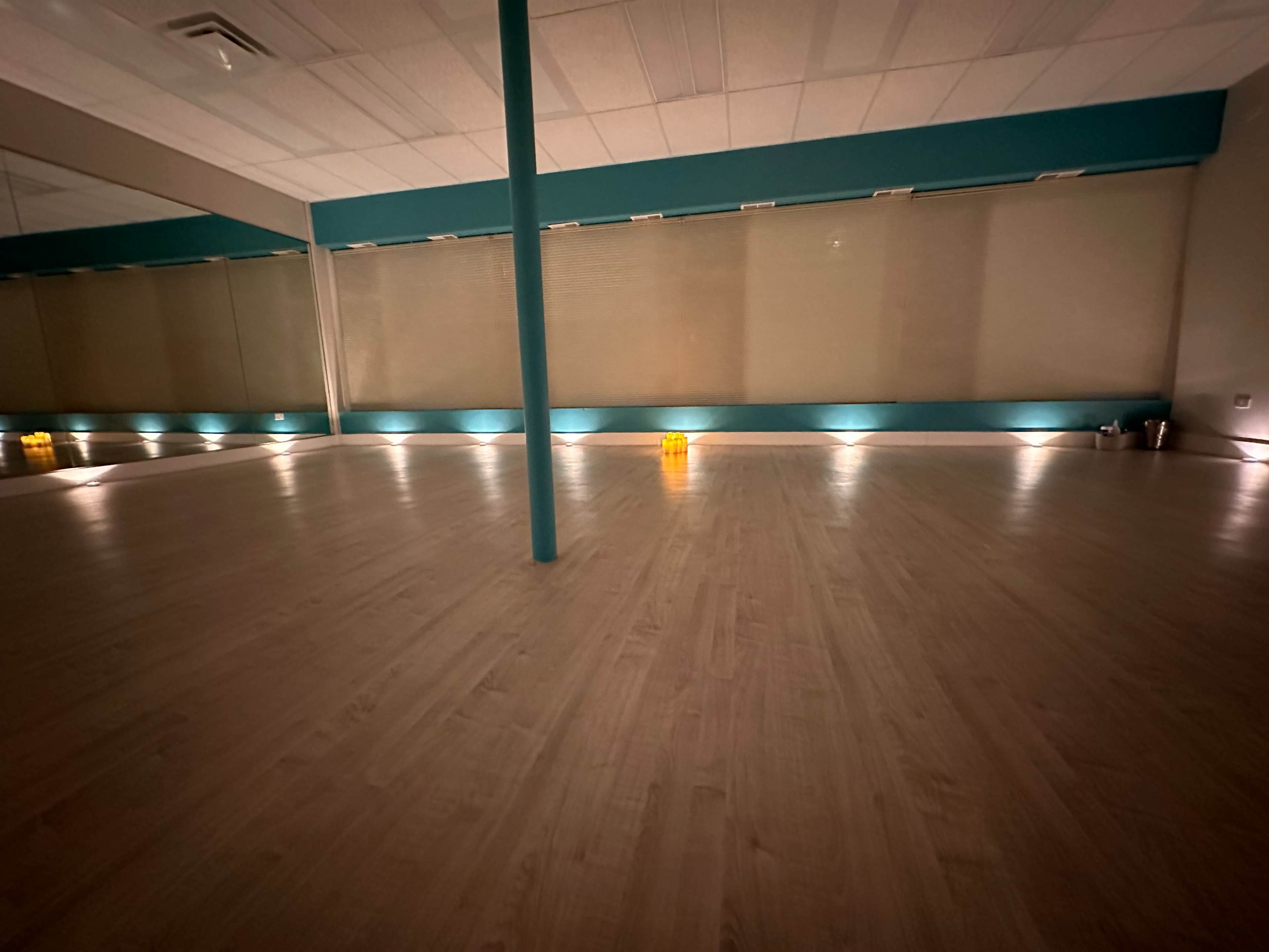 The image shows a spacious, empty room with wooden flooring, a central column, and soft lighting along the walls.