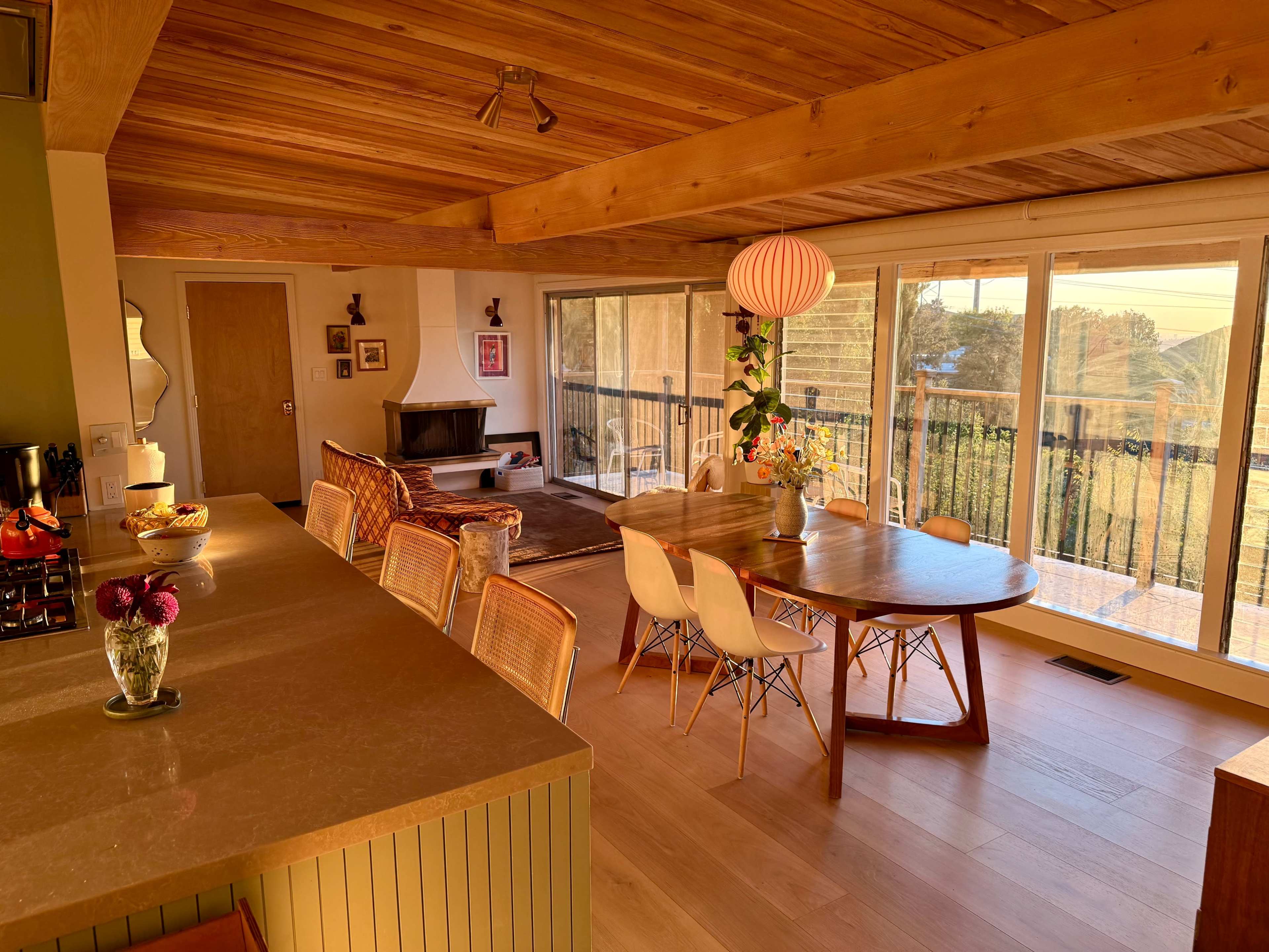 The image shows a modern interior space featuring a dining area with a round table, wooden chairs, and large windows that provide a view of the outdoors.