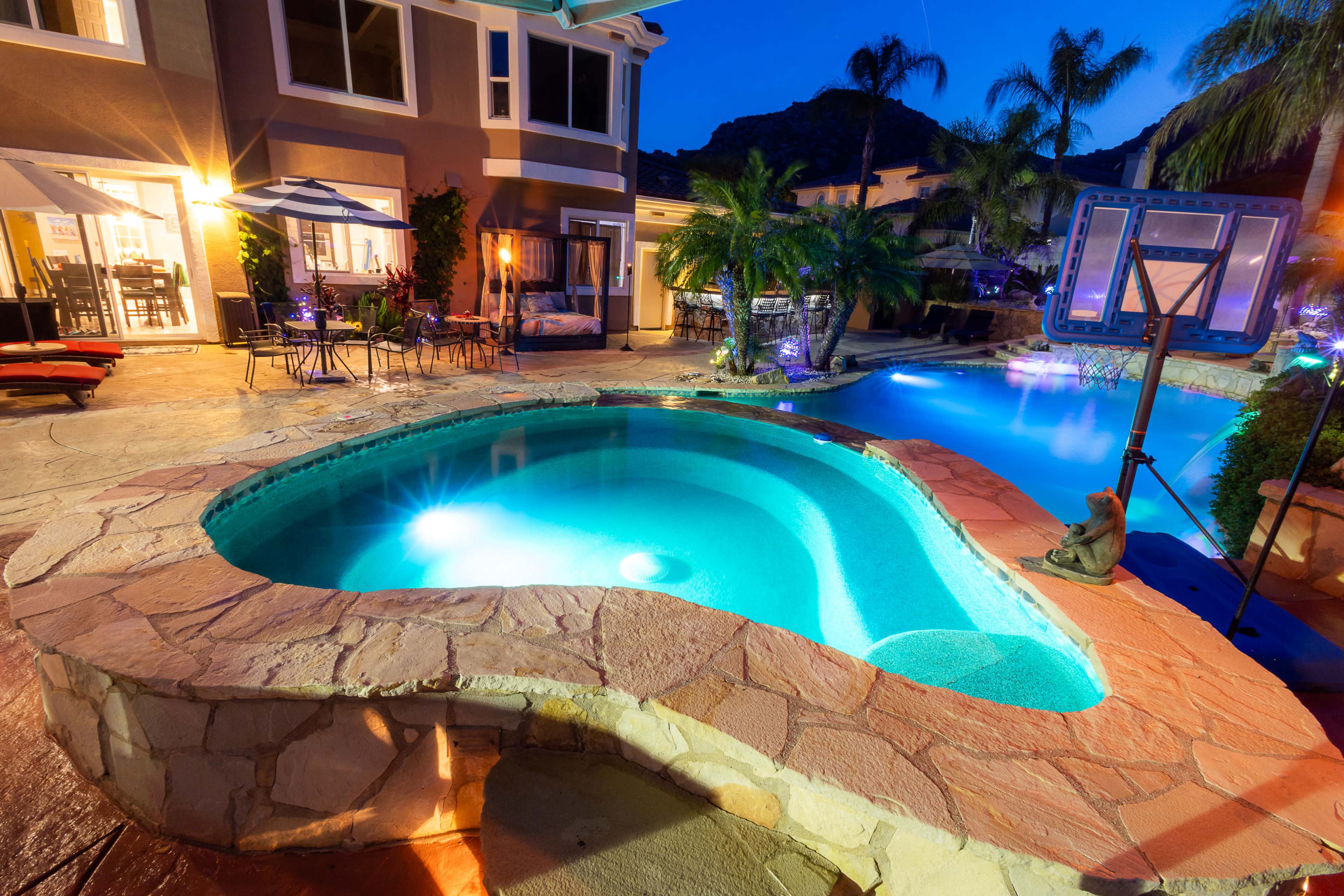 A backyard features a curved stone pool with a lounge area, surrounded by palm trees and illuminated by soft lighting.