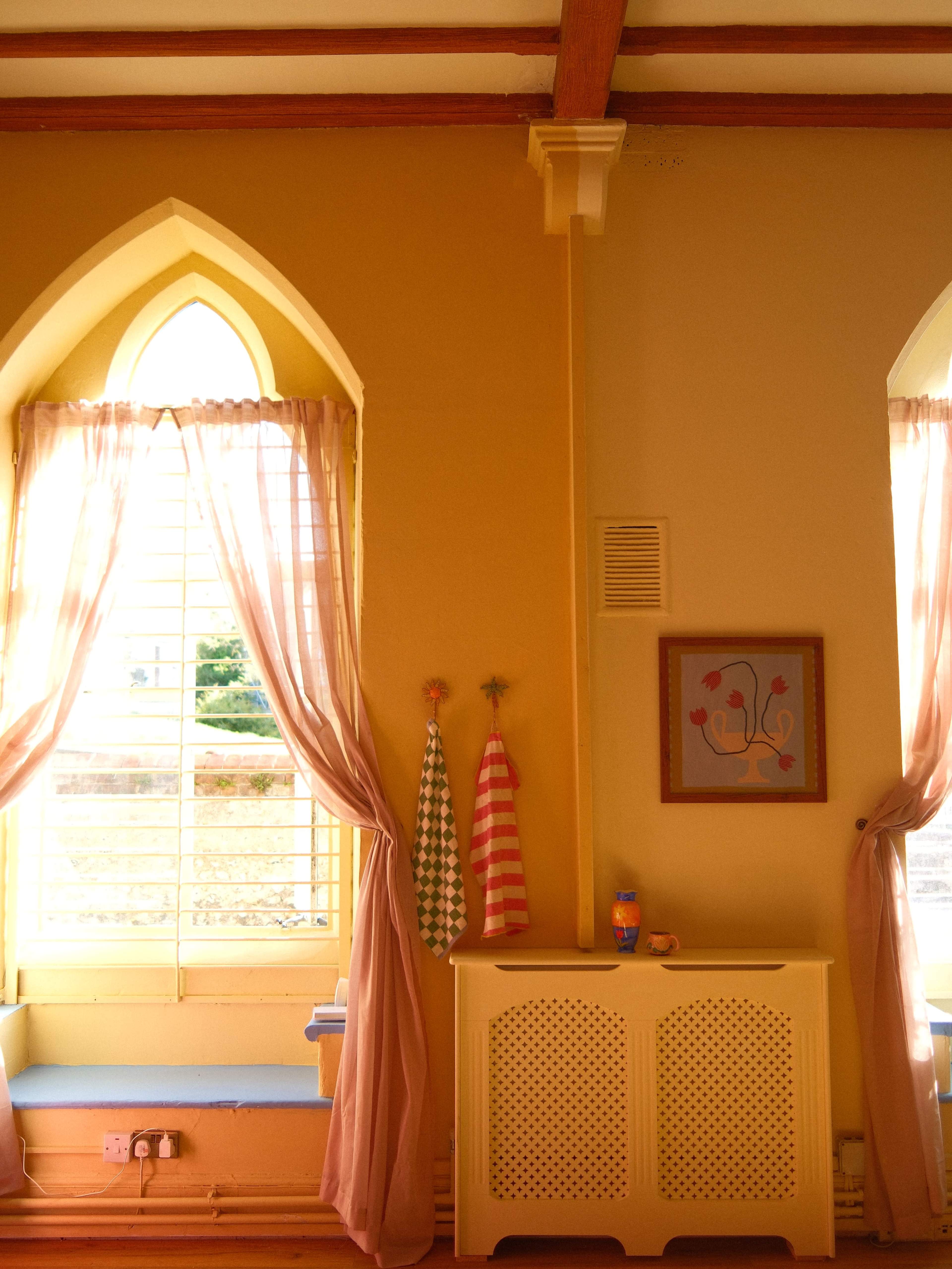 The image shows a room with yellow walls, a large arched window adorned with curtains, and decorative items hanging on the wall.
