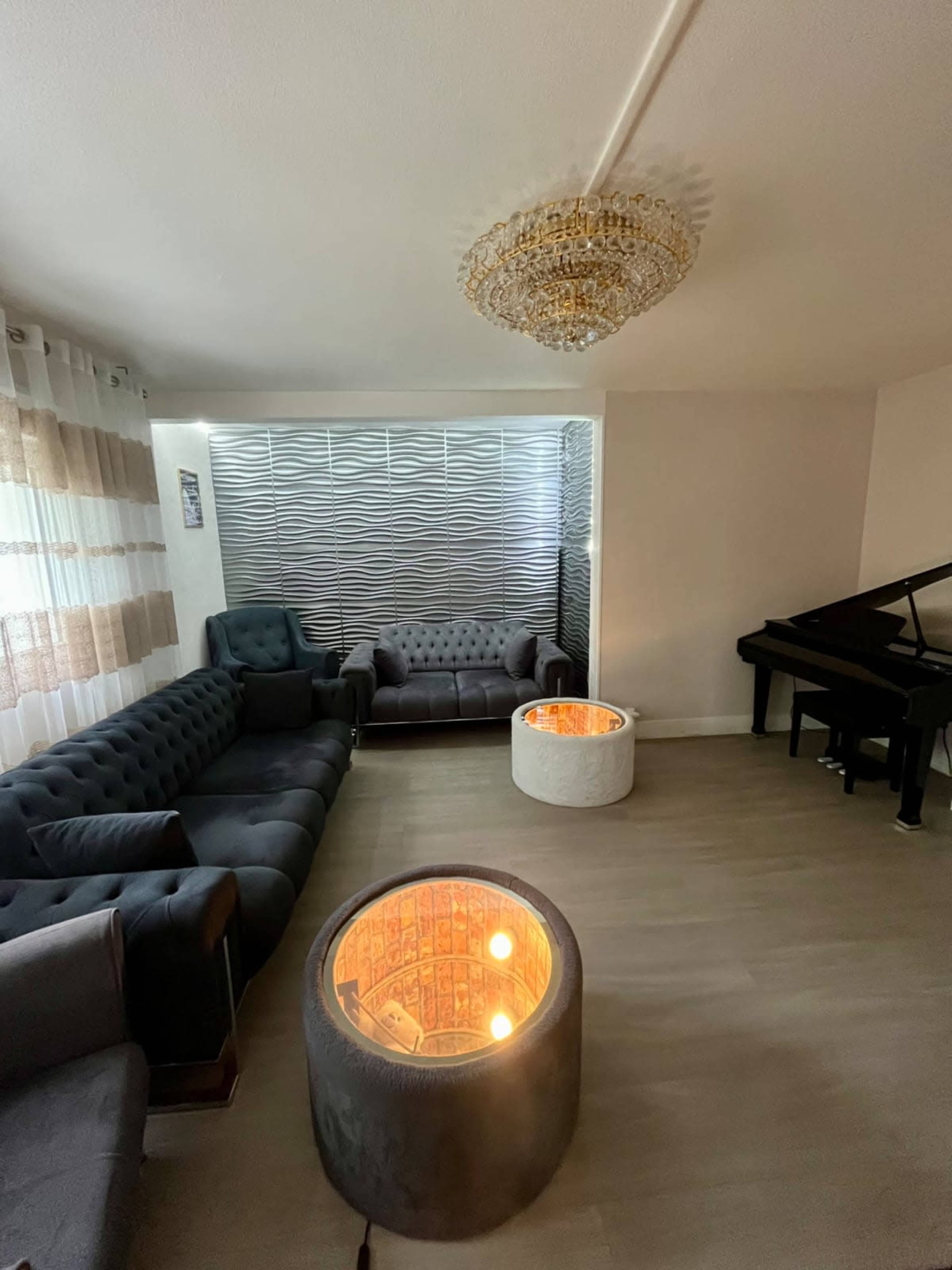The image depicts a modern living room with gray furniture, including a sectional sofa, a piano, and two round illuminated tables.
