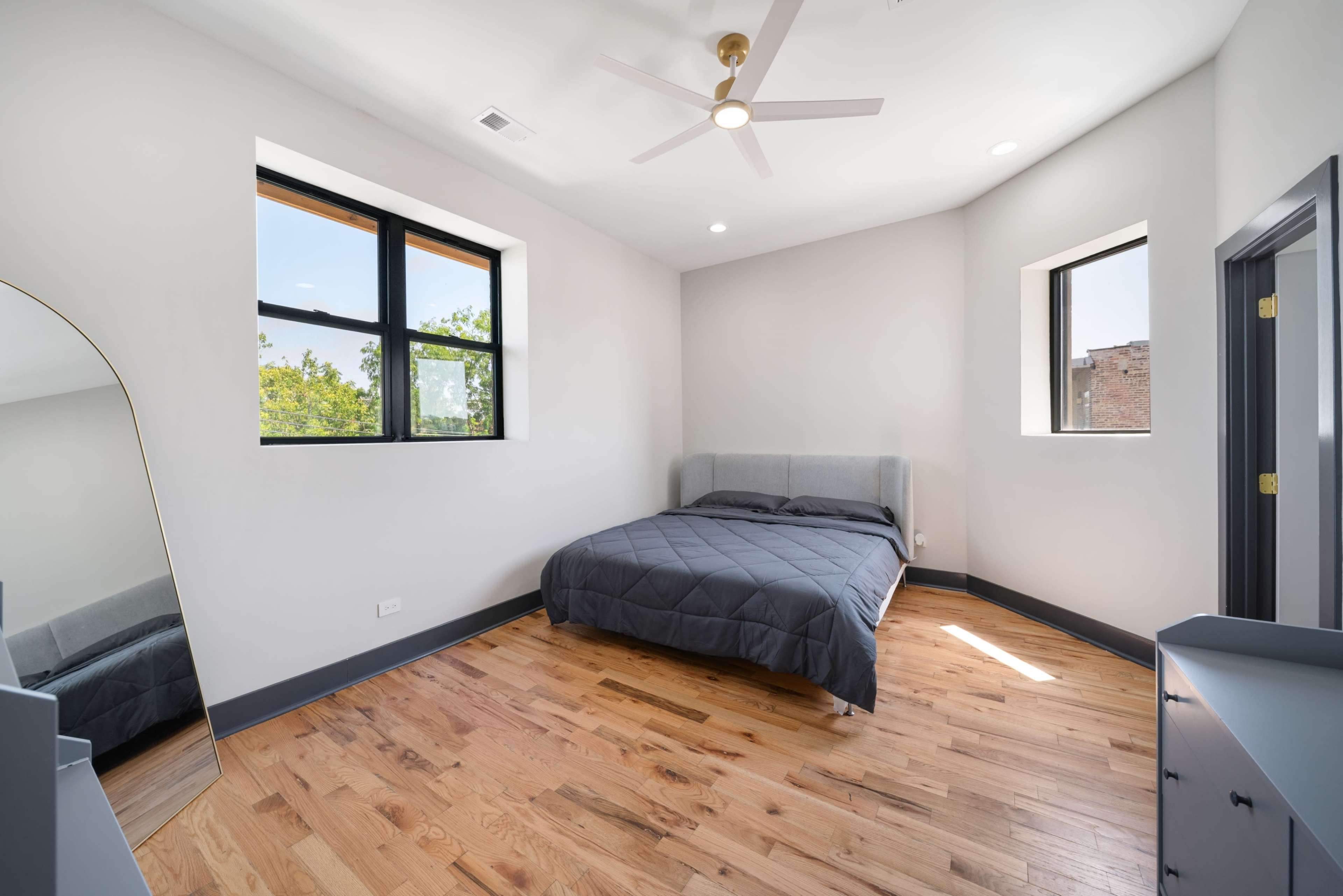 The image shows a bedroom with a bed, two windows, a mirror, and a drawer unit, all set against a wood floor.