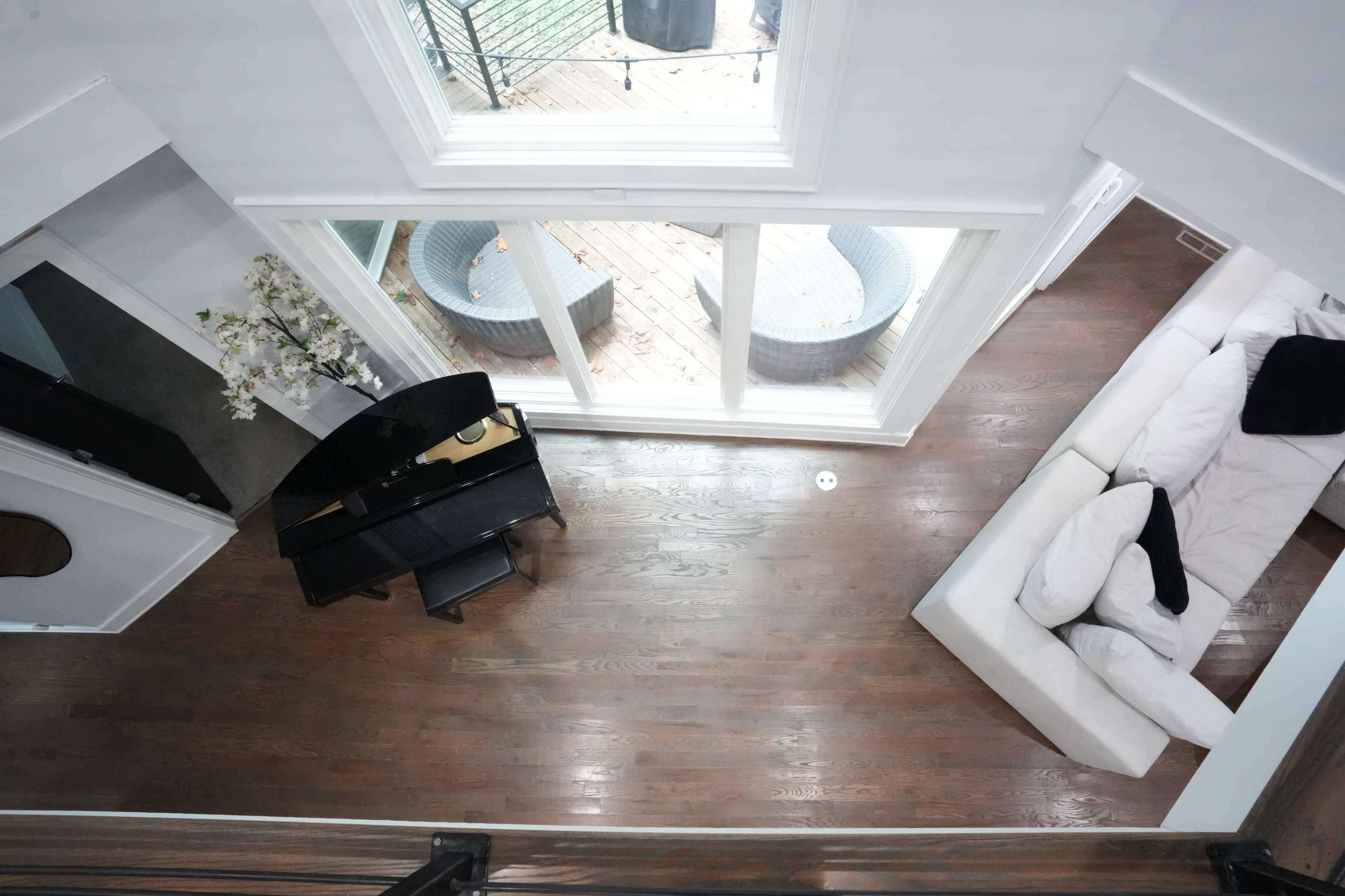 The image shows a spacious living area viewed from above, featuring a black piano, a white sectional sofa, and large windows that open to a deck.