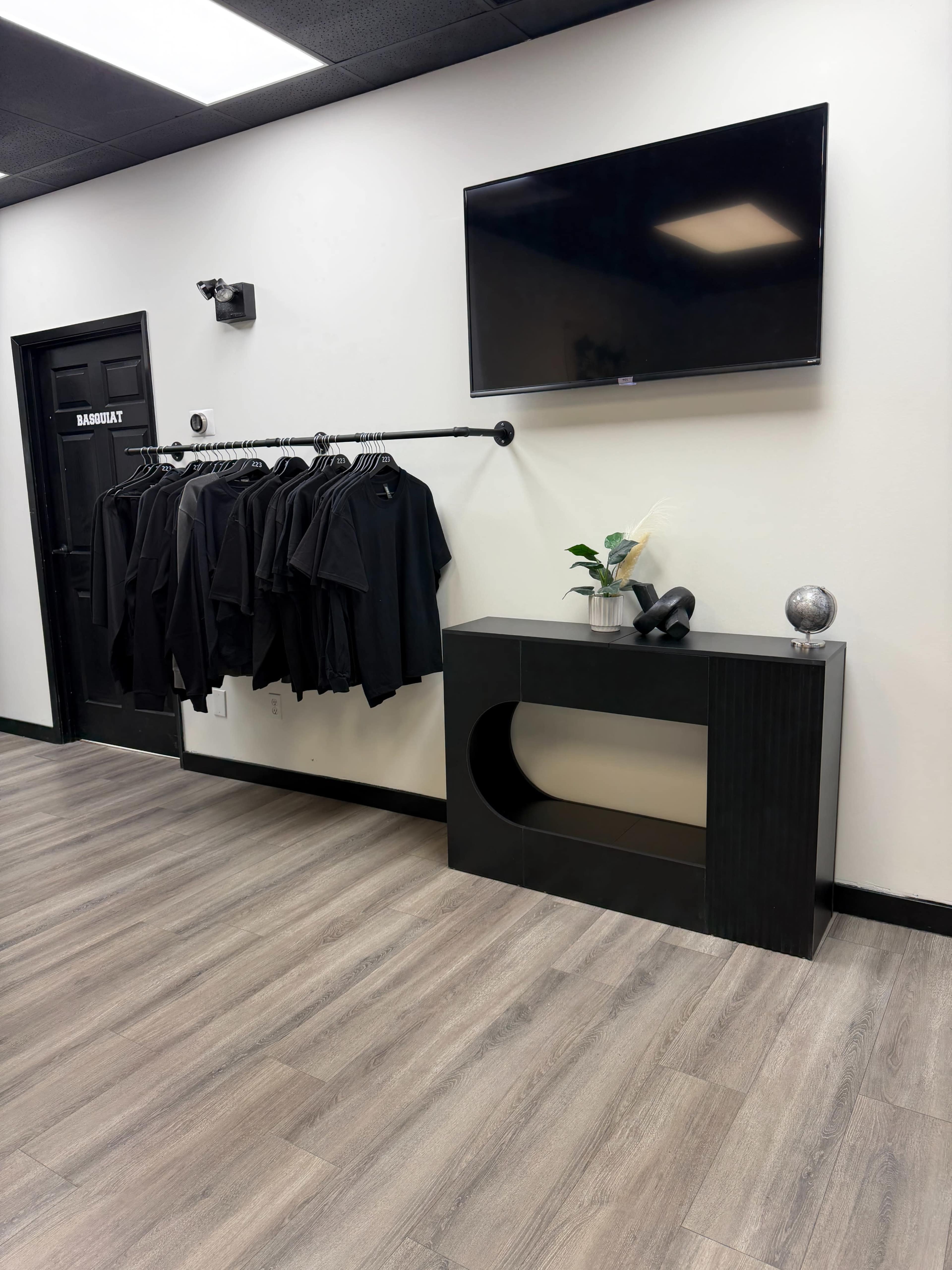The image shows a room with a black clothing rack holding several black garments, a console table beneath a television, and a plant and decorative globe on the table.
