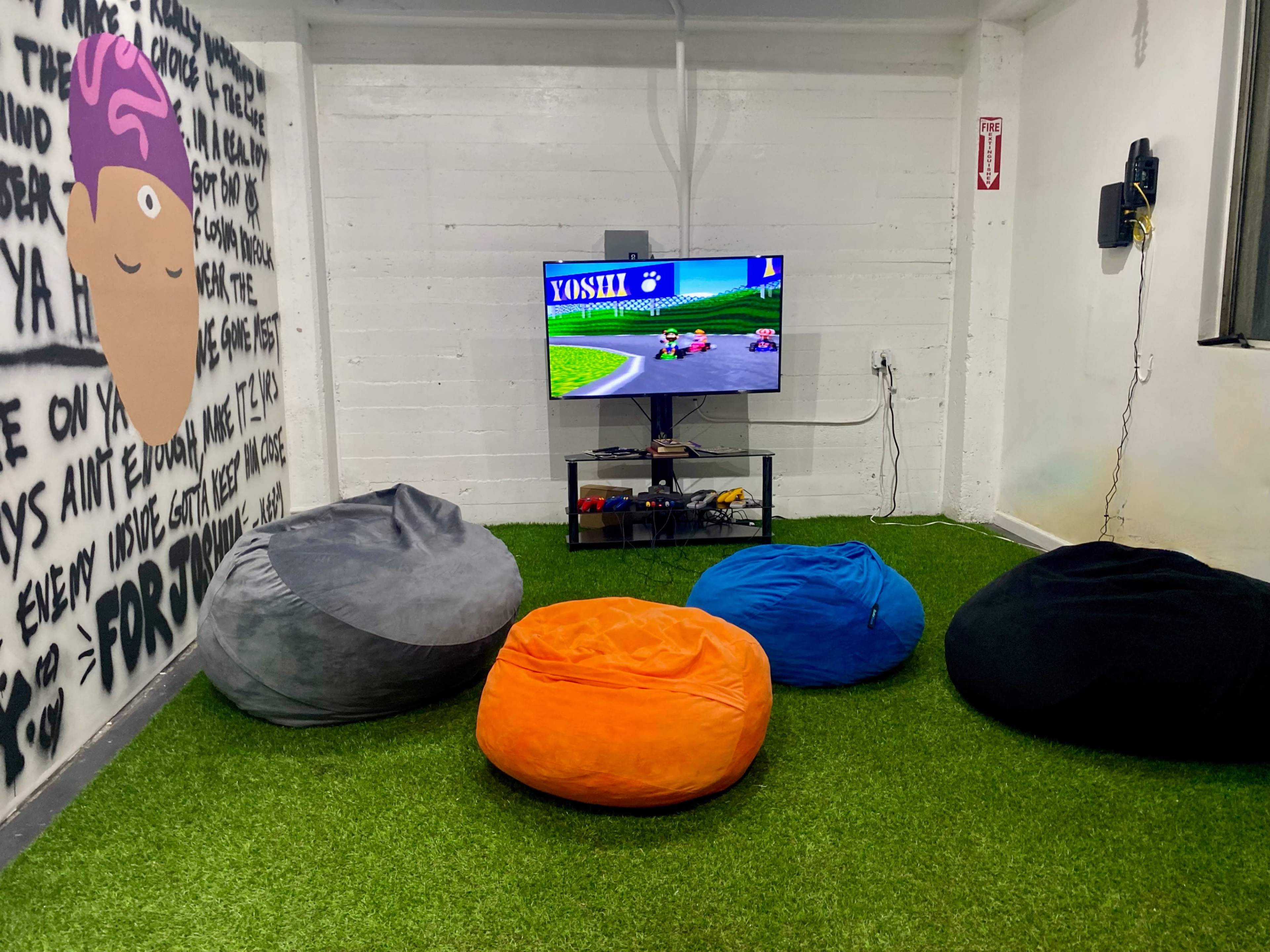 The image shows a small room with a television displaying a racing game, surrounded by several bean bag chairs on artificial grass.