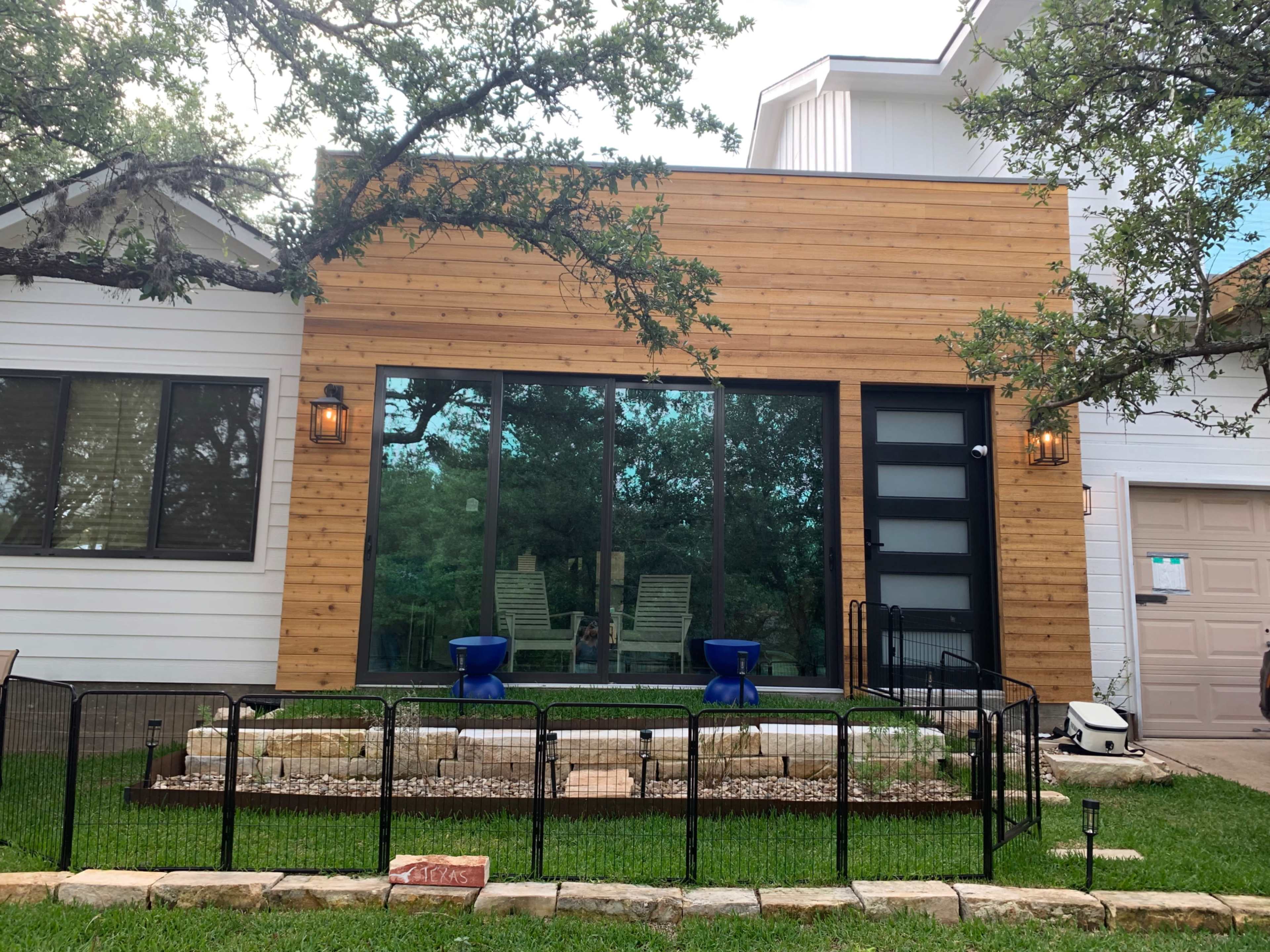 A modern home exterior featuring a wooden facade, large glass windows, and a fenced front yard with stone landscaping.