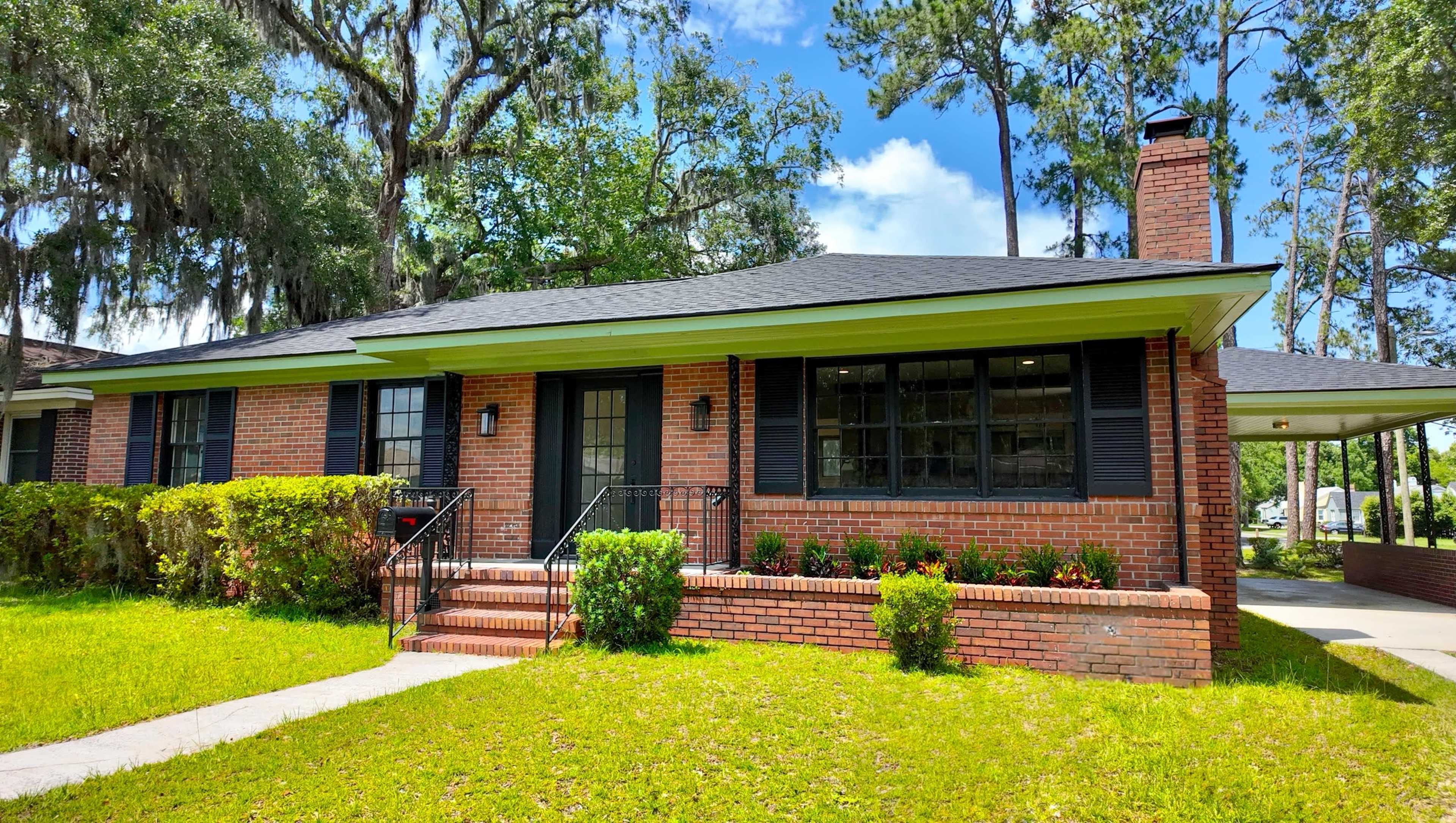 The image shows a brick single-story house with a green trim, a front porch, and a well-maintained lawn surrounded by trees.
