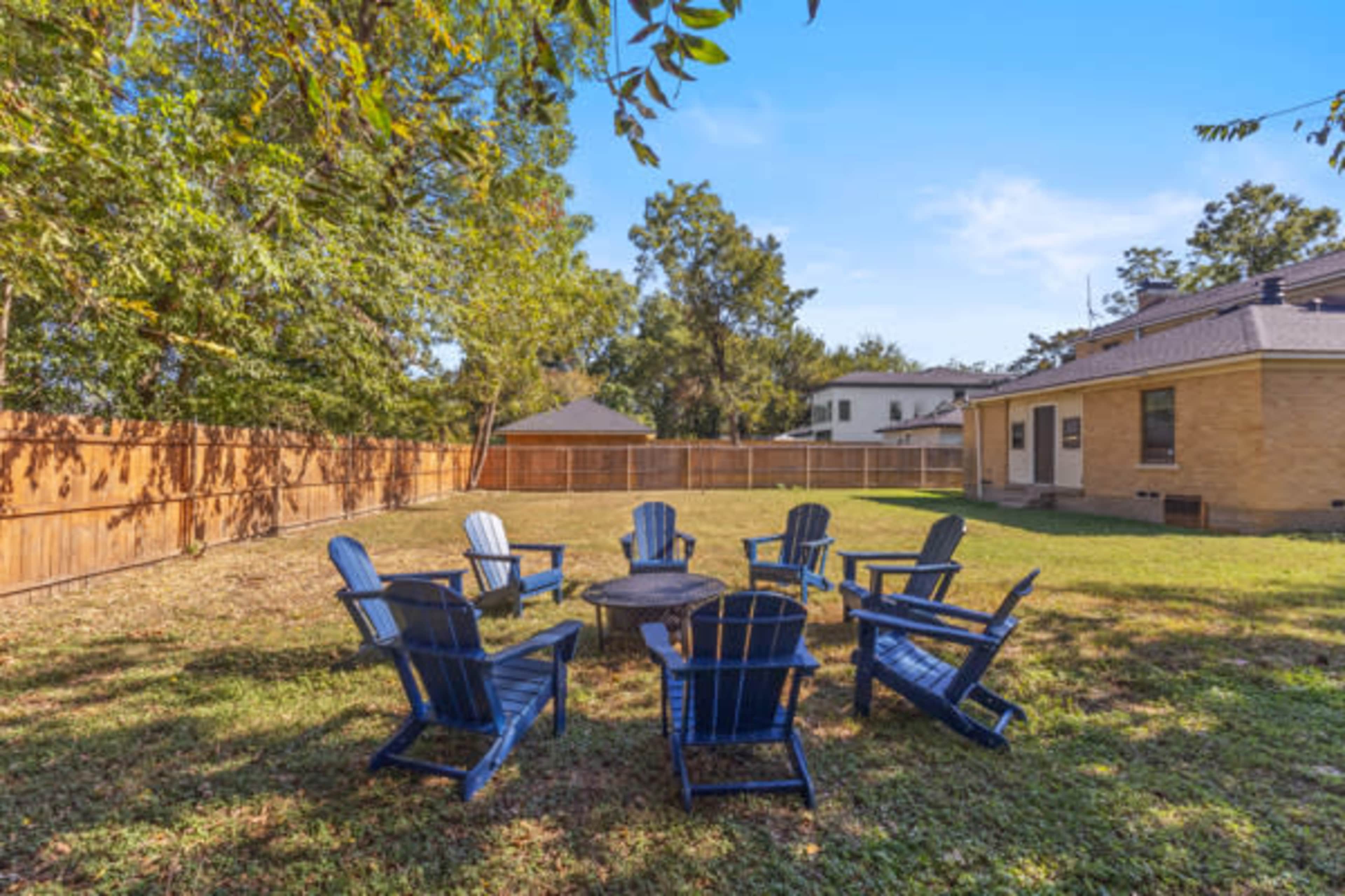The image shows a backyard with a circular arrangement of blue Adirondack chairs surrounding a fire pit.