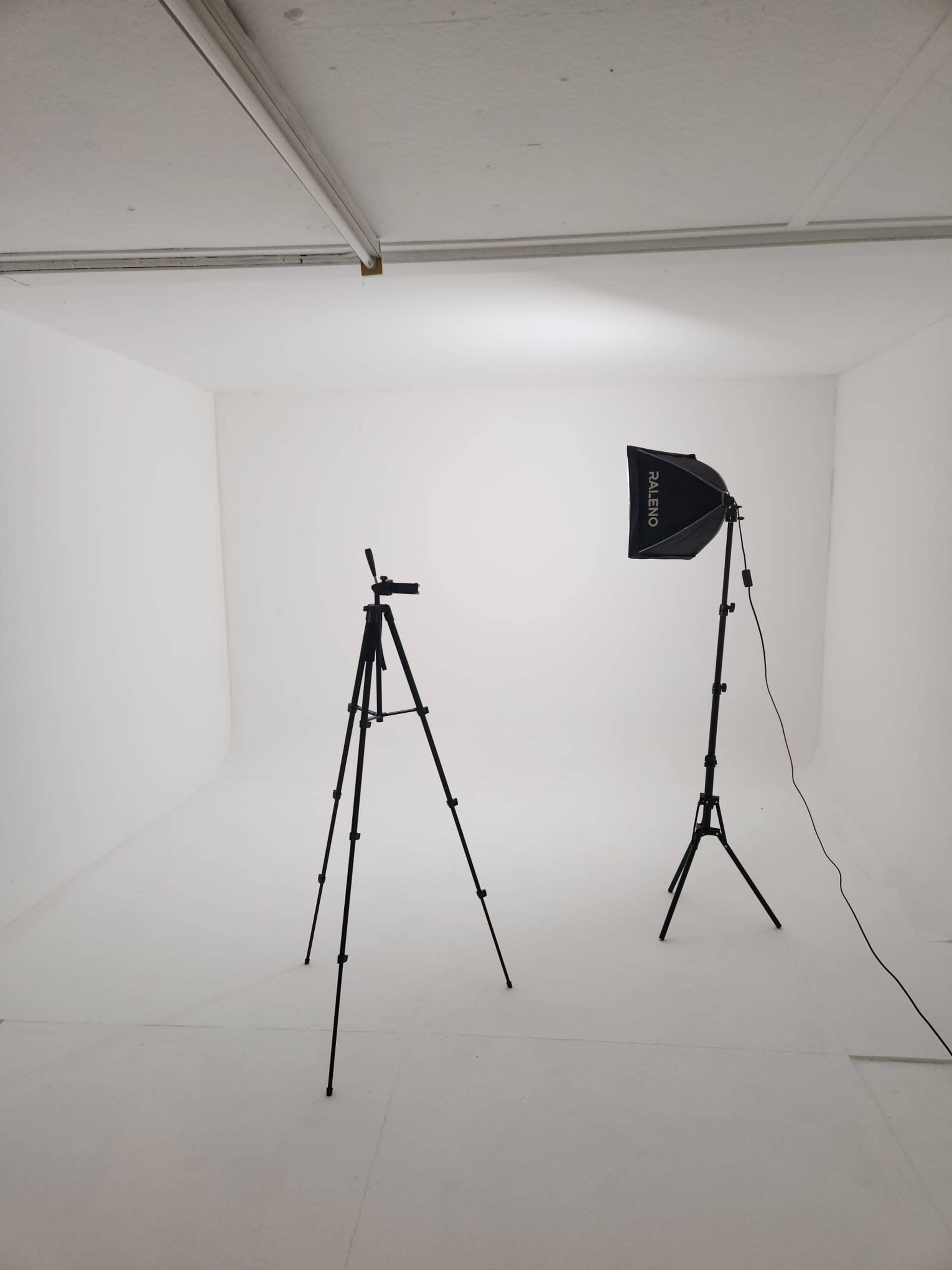 The image shows a minimal photography studio setup featuring a tripod and a softbox light in a white room.