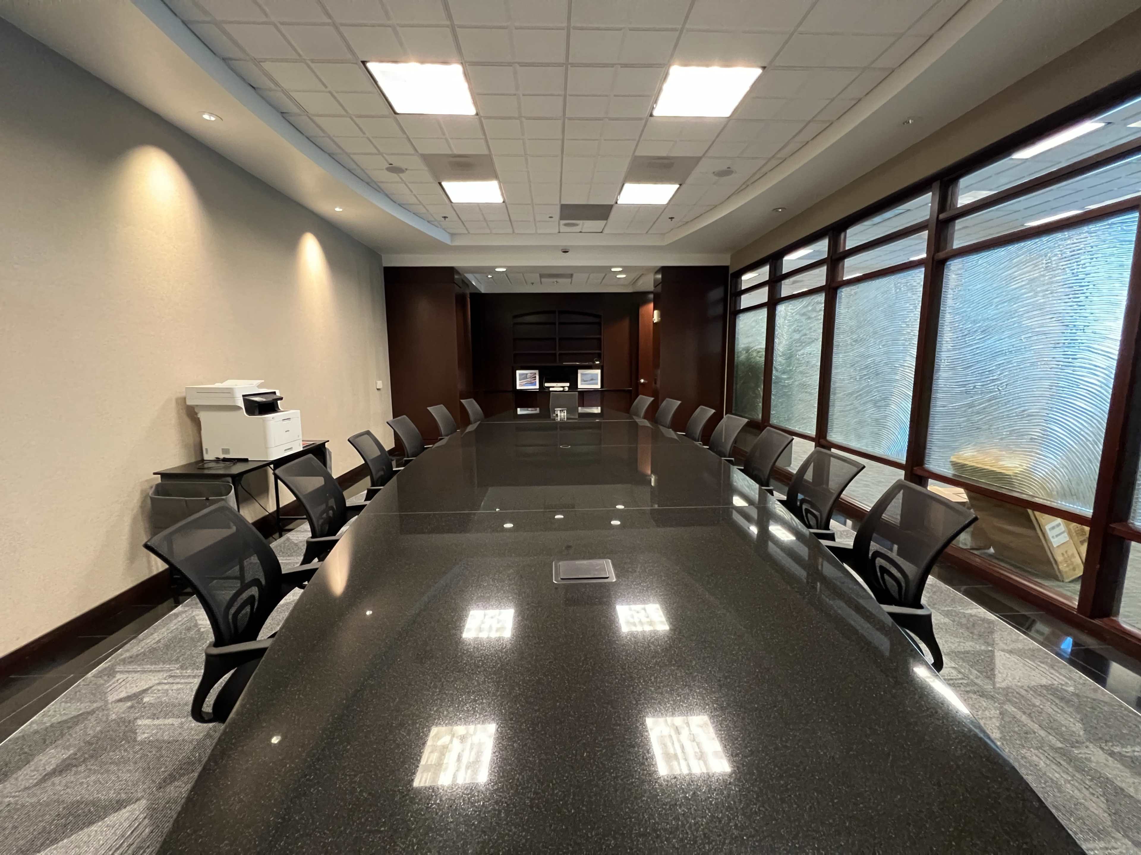 A large, empty conference room features a long table surrounded by ergonomic chairs, with a printer and glass windows along one wall.