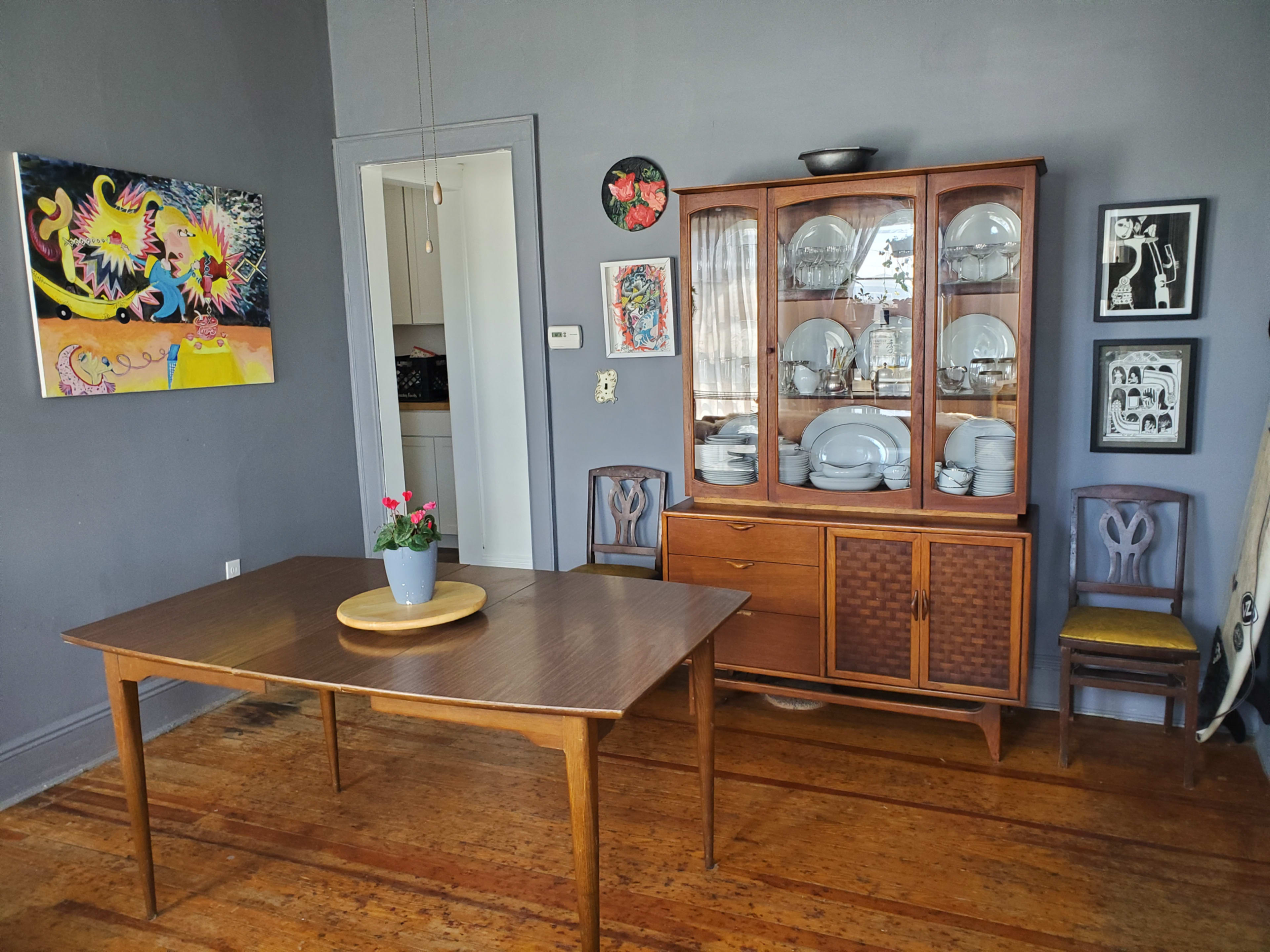 A wooden dining table is positioned in a room with gray walls, alongside a glass-fronted cabinet filled with dishware and various pieces of artwork displayed on the walls.