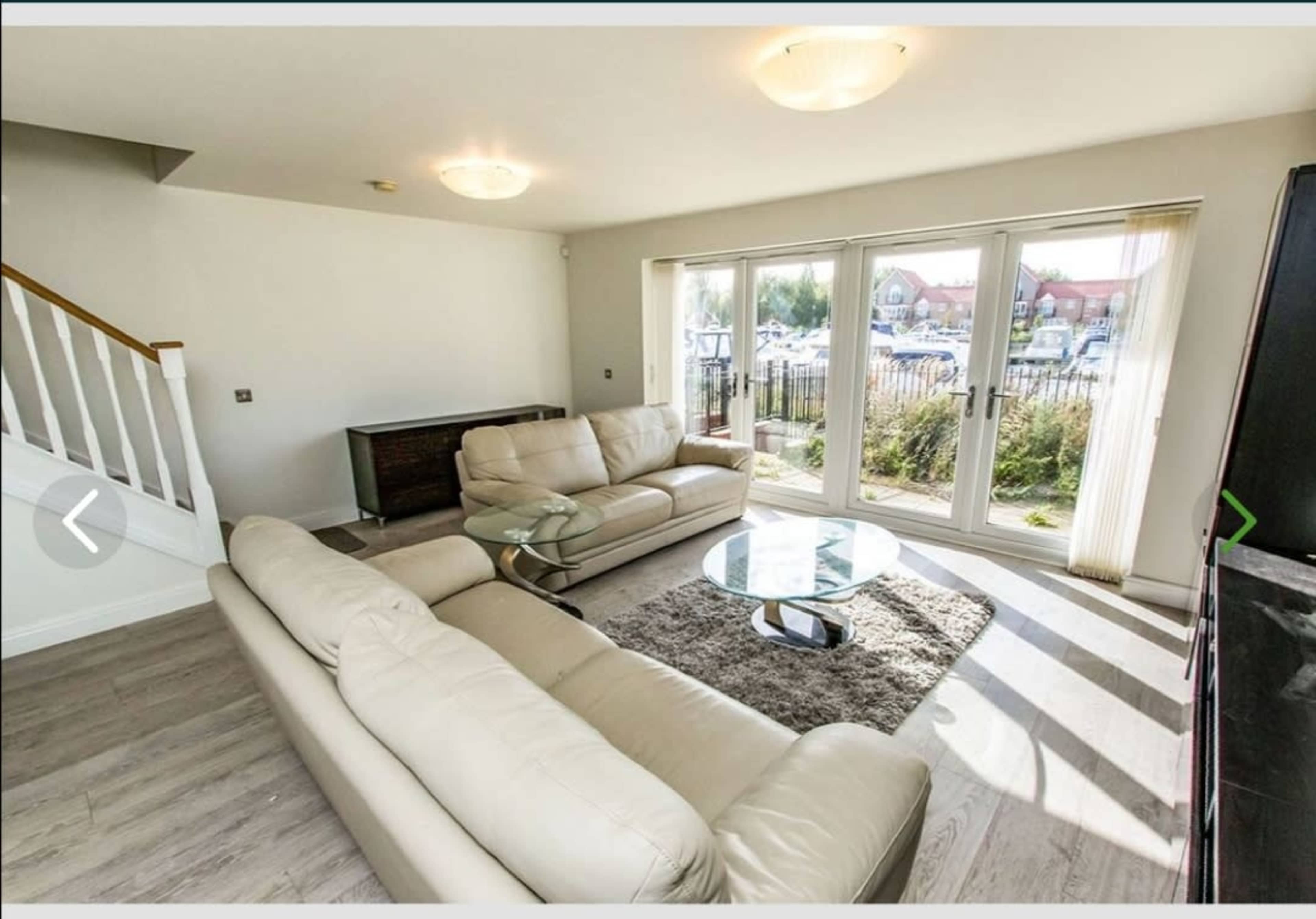 The image shows a living room with a light-colored sectional sofa, a glass coffee table on a rug, and large windows leading to a patio area.