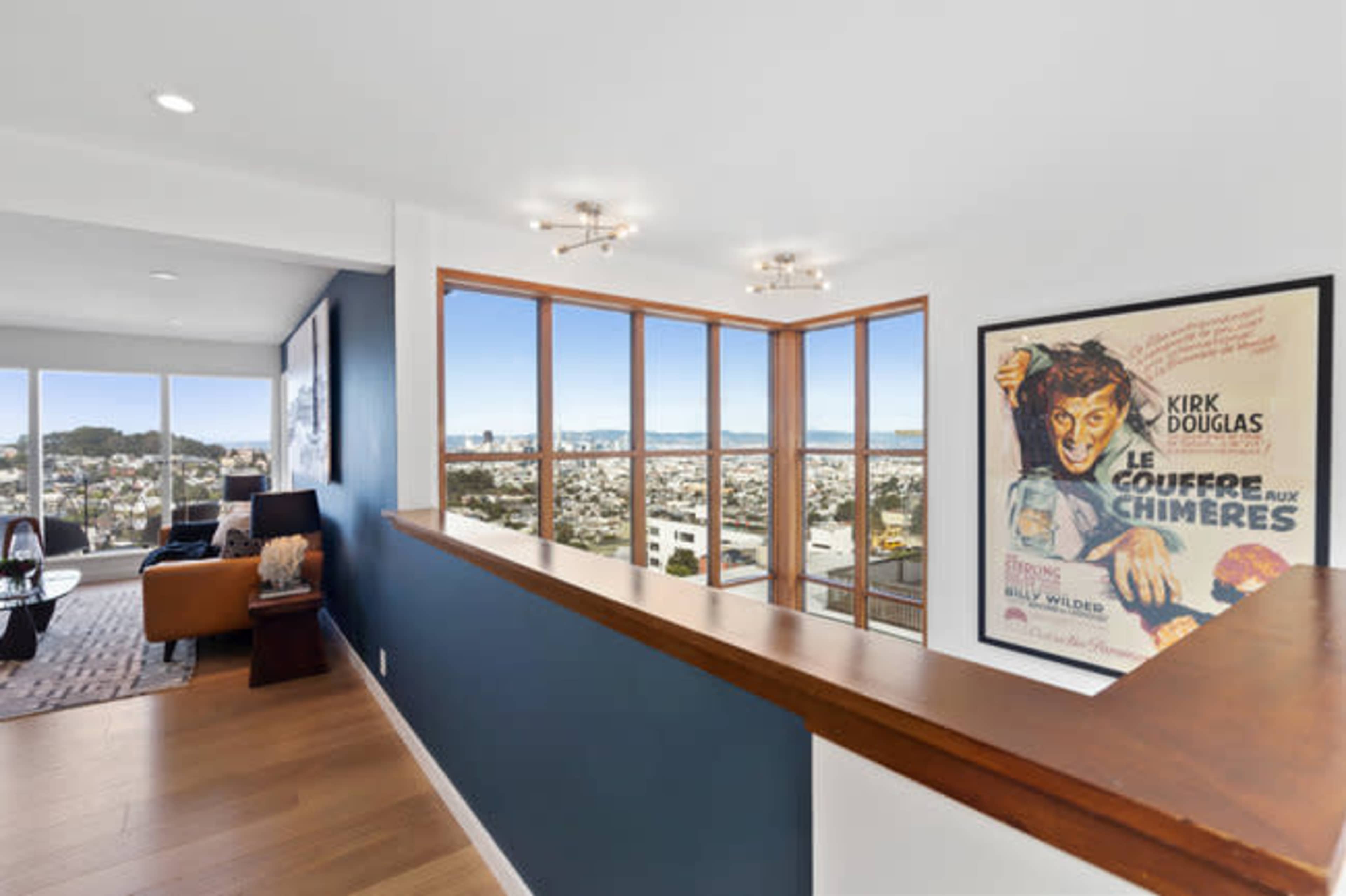 A modern hallway features a wooden railing, large windows with city views, and a vintage movie poster on the wall.