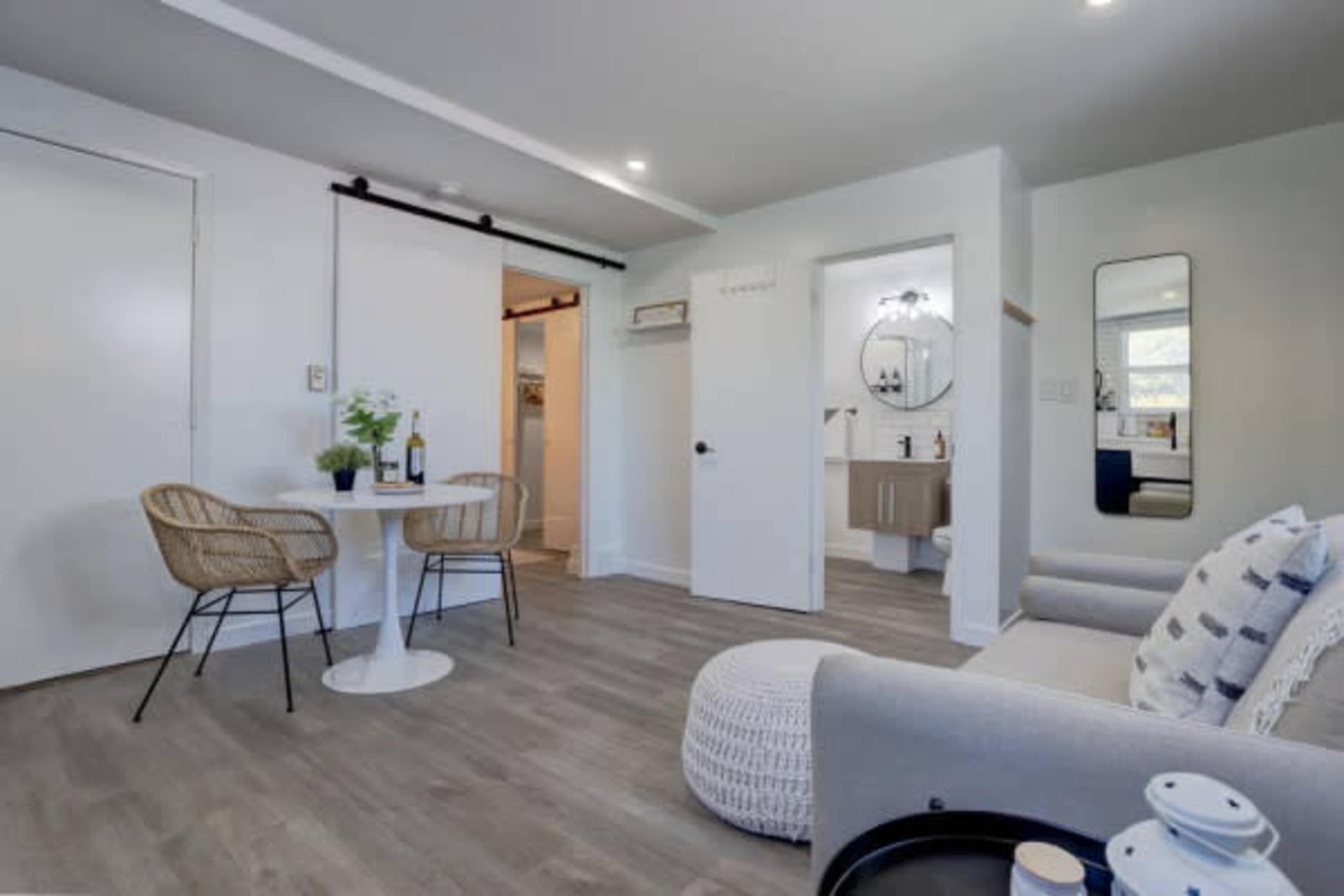 A modern, minimalistic living space featuring a small table with two chairs, a sofa, and a bathroom door in a well-lit room.