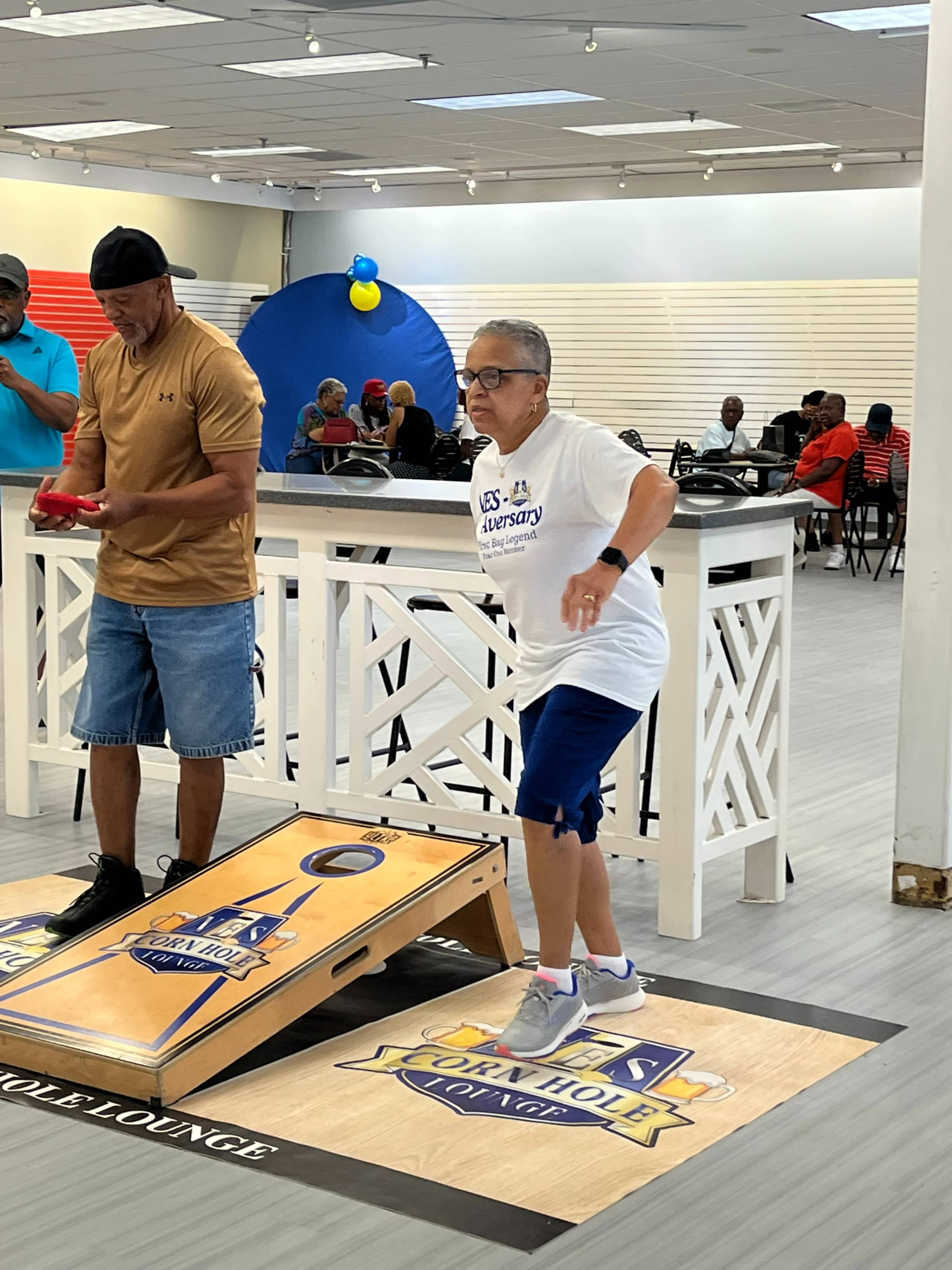 A person in a white T-shirt is throwing a bean bag toward a cornhole board while others participate in the game in the background.