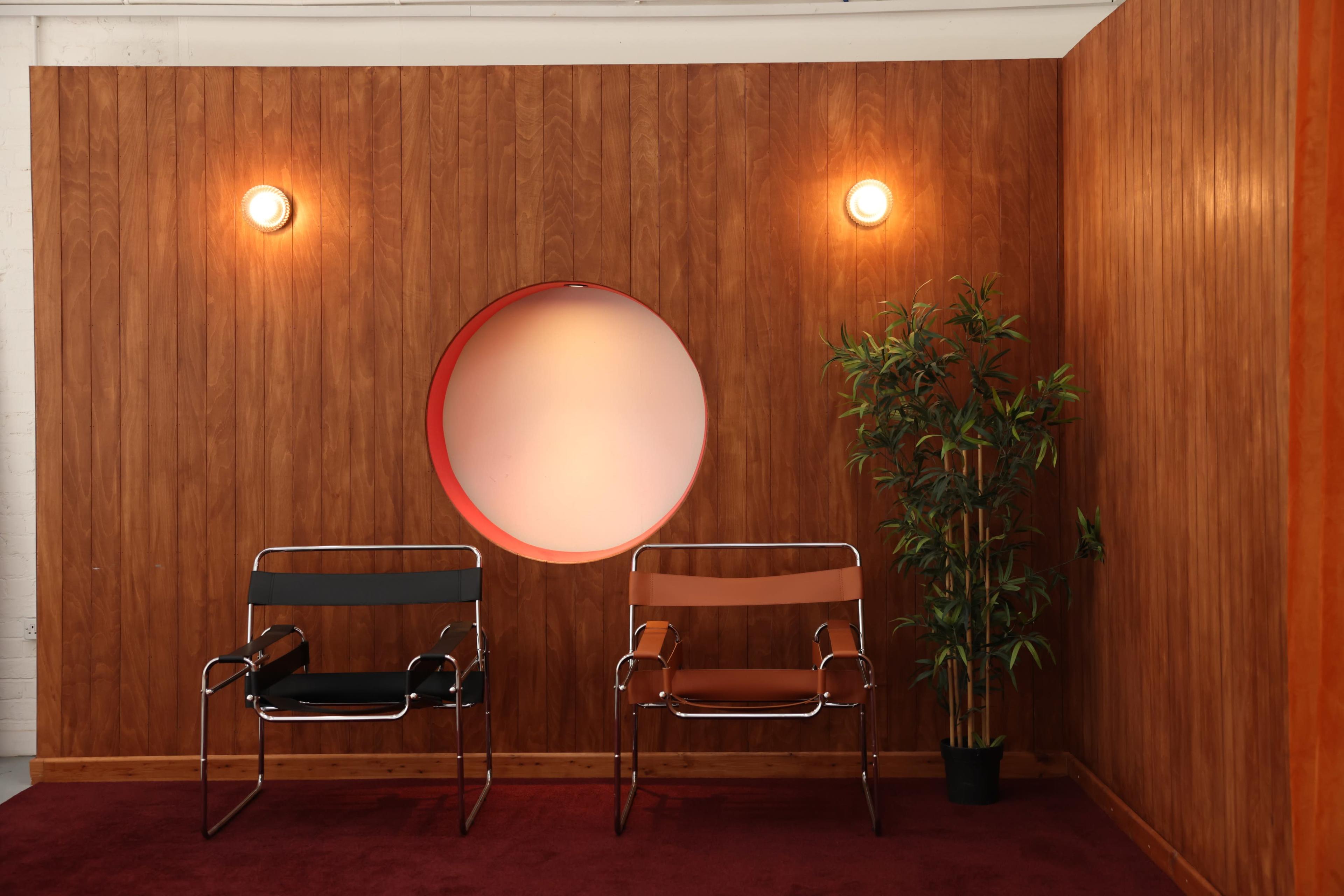 The image shows two modern chairs placed against a wooden wall with a circular cutout, accompanied by a potted plant.