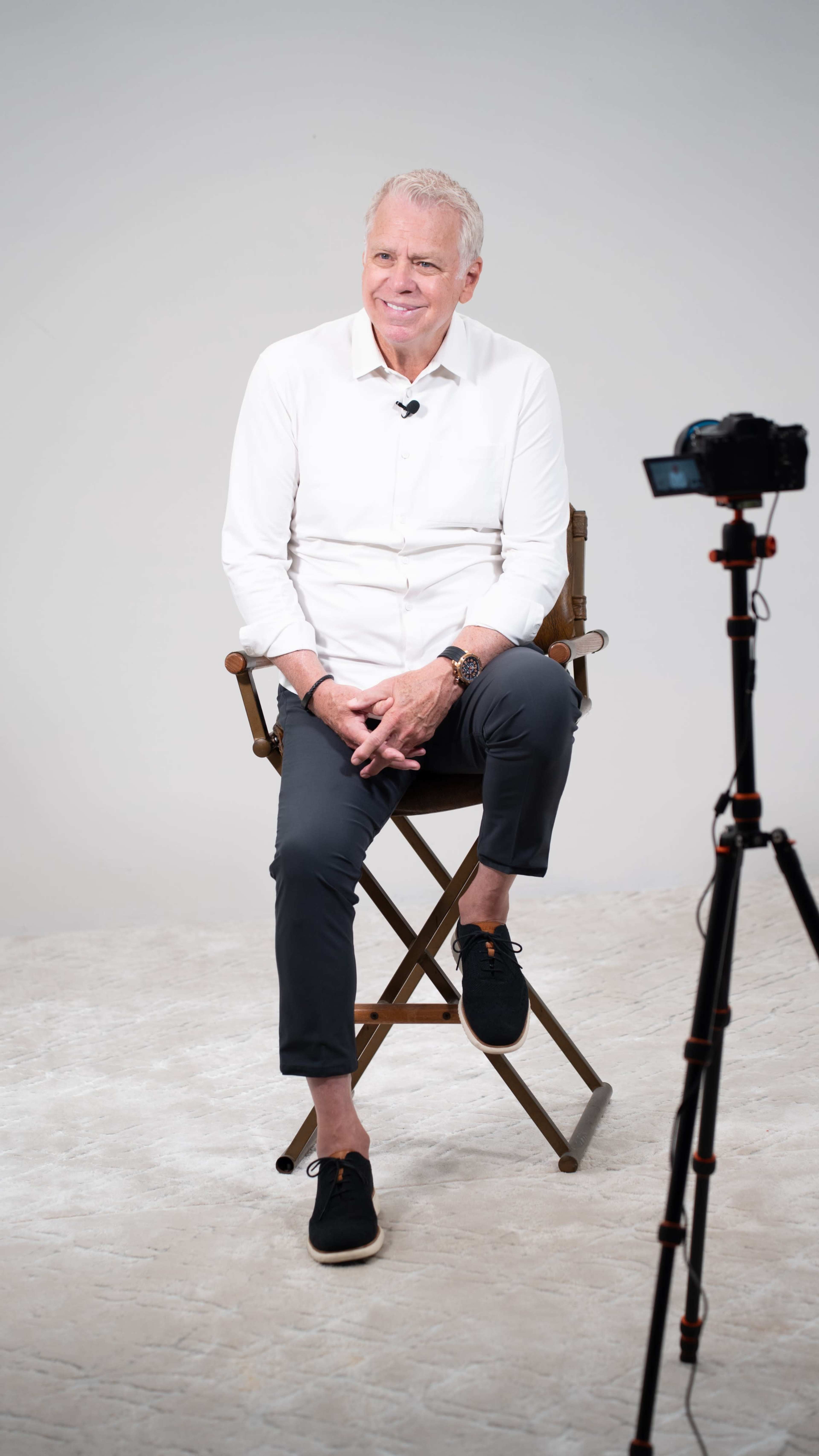 A man in a white shirt and dark pants sits on a wooden stool in front of a camera setup against a plain background.