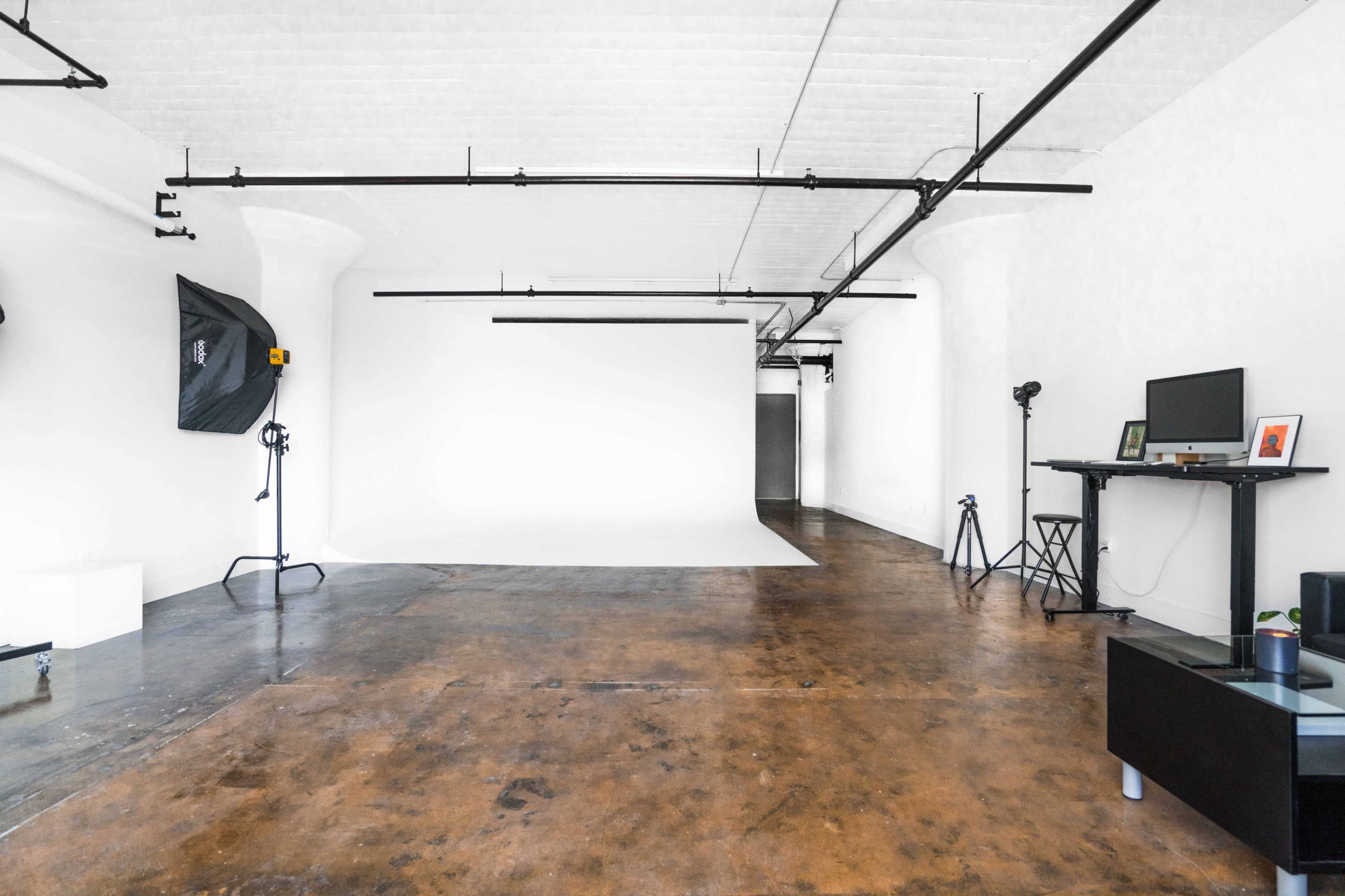 A spacious, empty photography studio with a white backdrop, metal supports, adjustable lighting equipment, and a minimalist setup.