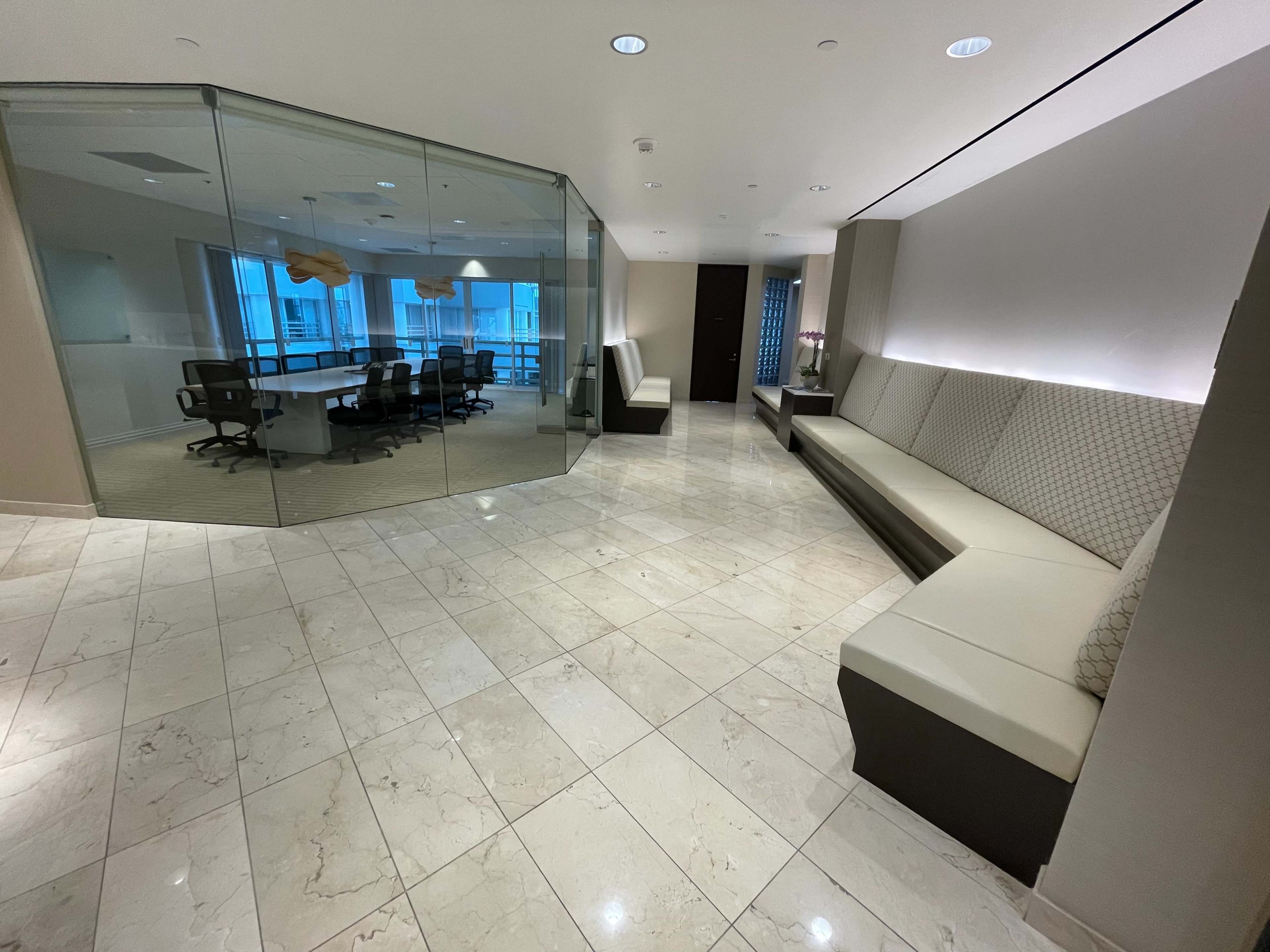 An office lobby features tiled flooring, sleek seating along the walls, and a glass-walled meeting room in the background.