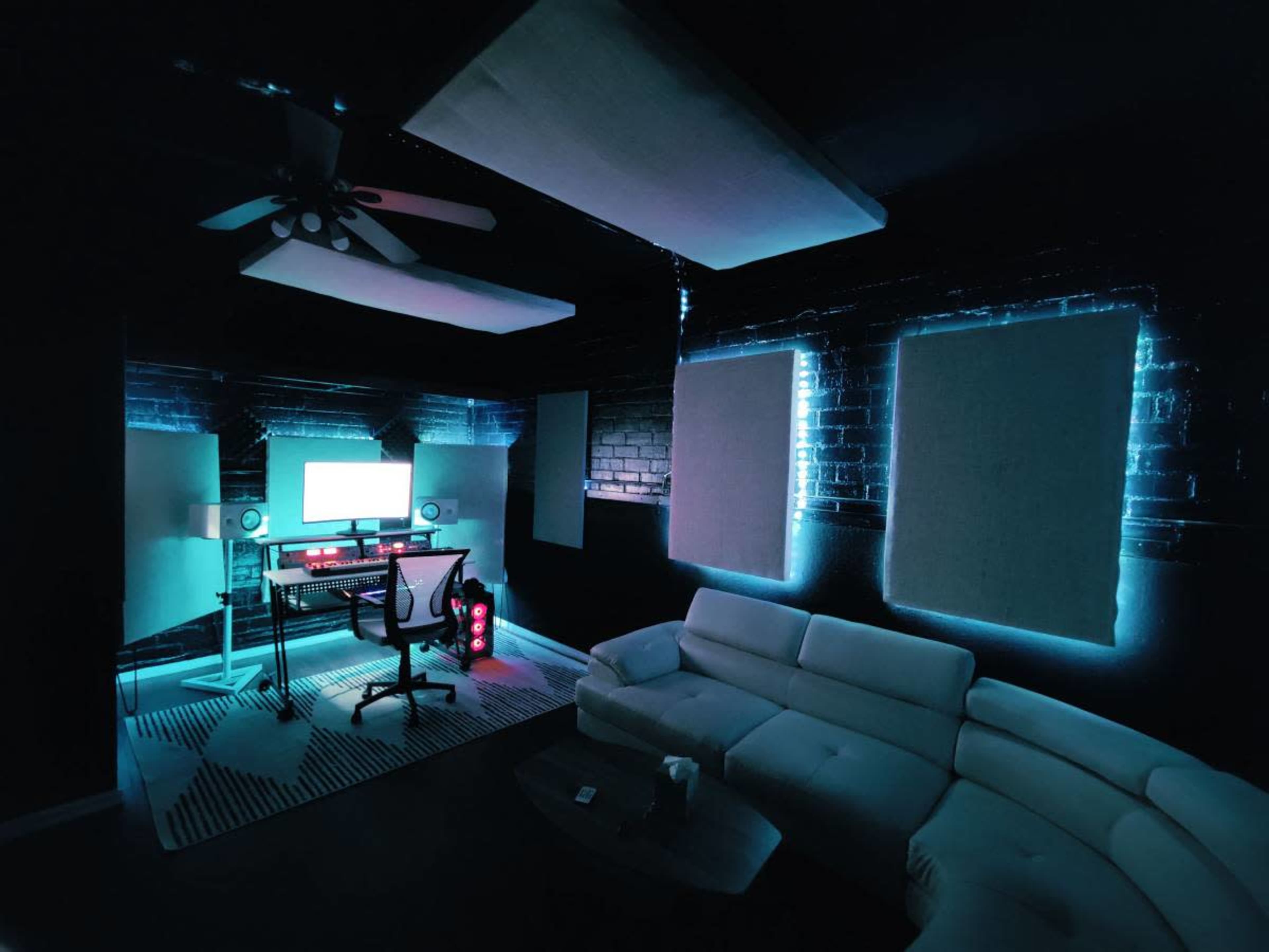 A modern music studio features a white curved sofa, a sleek desk with a computer setup, and soundproofing panels illuminated by soft blue lighting.