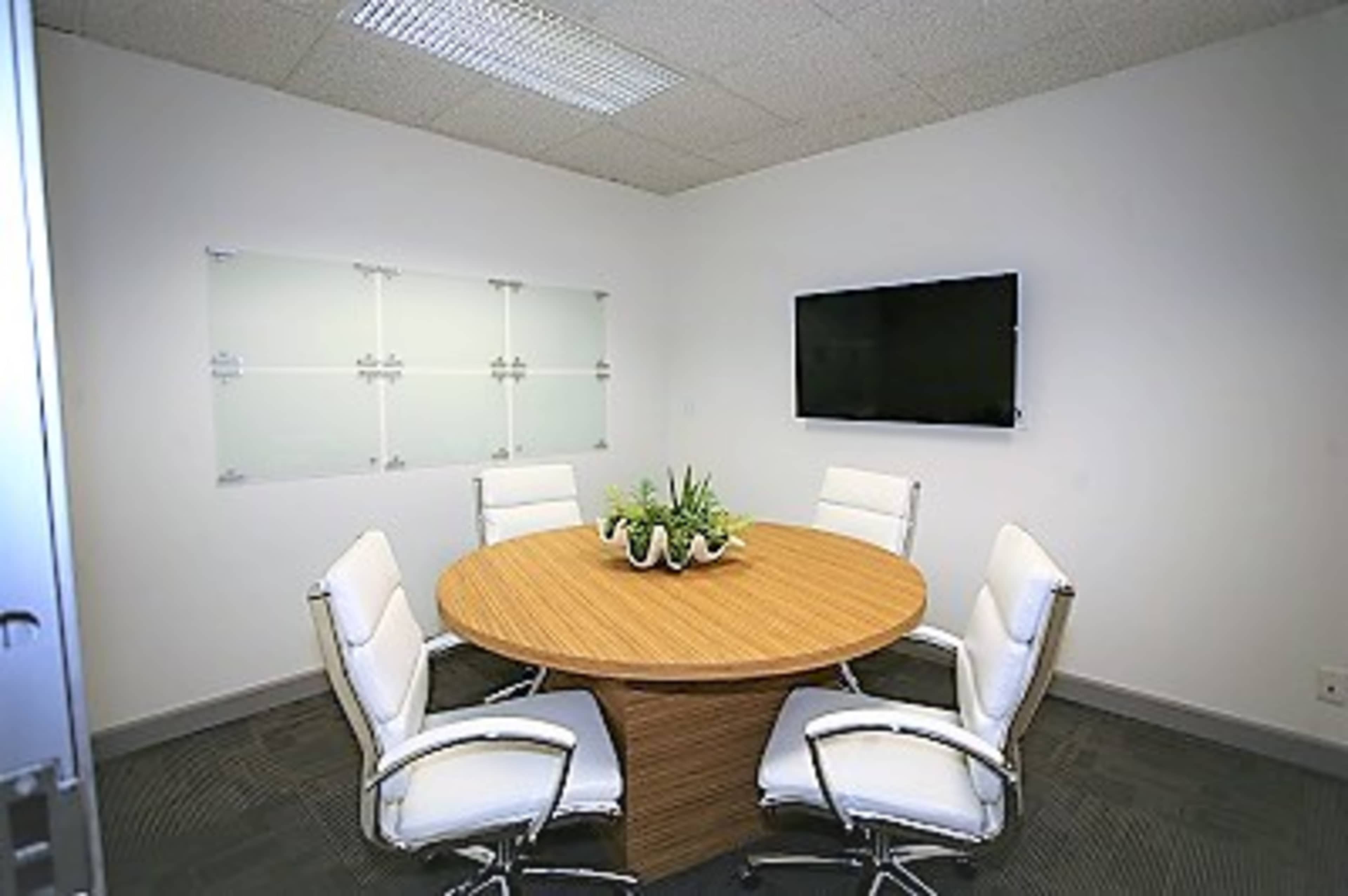 A conference room features a round wooden table surrounded by four white chairs, with a television mounted on the wall and a plant centerpieces on the table.