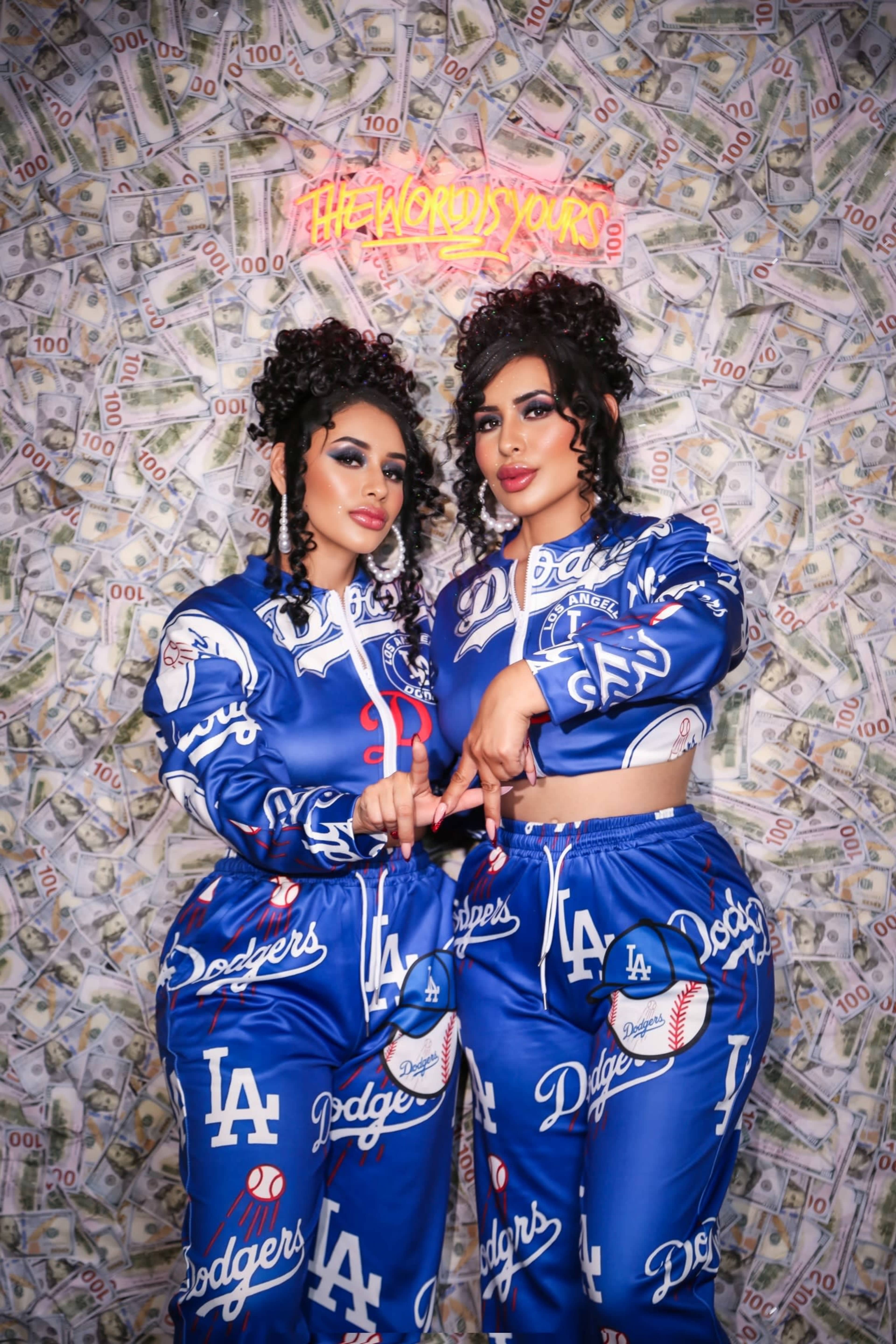 Two women pose in matching blue Los Angeles Dodgers outfits in front of a wall covered in money.