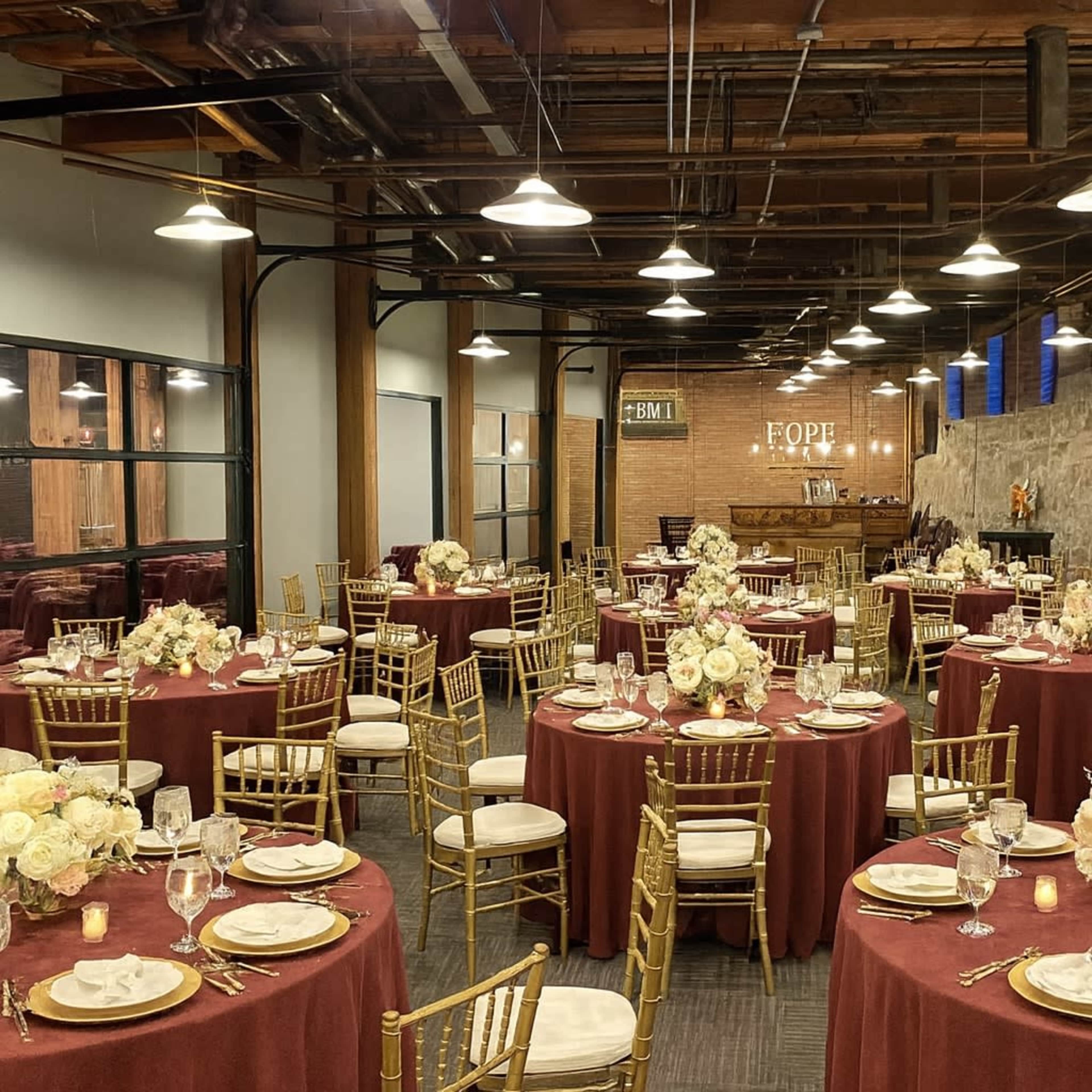 The image shows a banquet hall with circular tables dressed in burgundy tablecloths, adorned with floral centerpieces and surrounded by gold chairs.