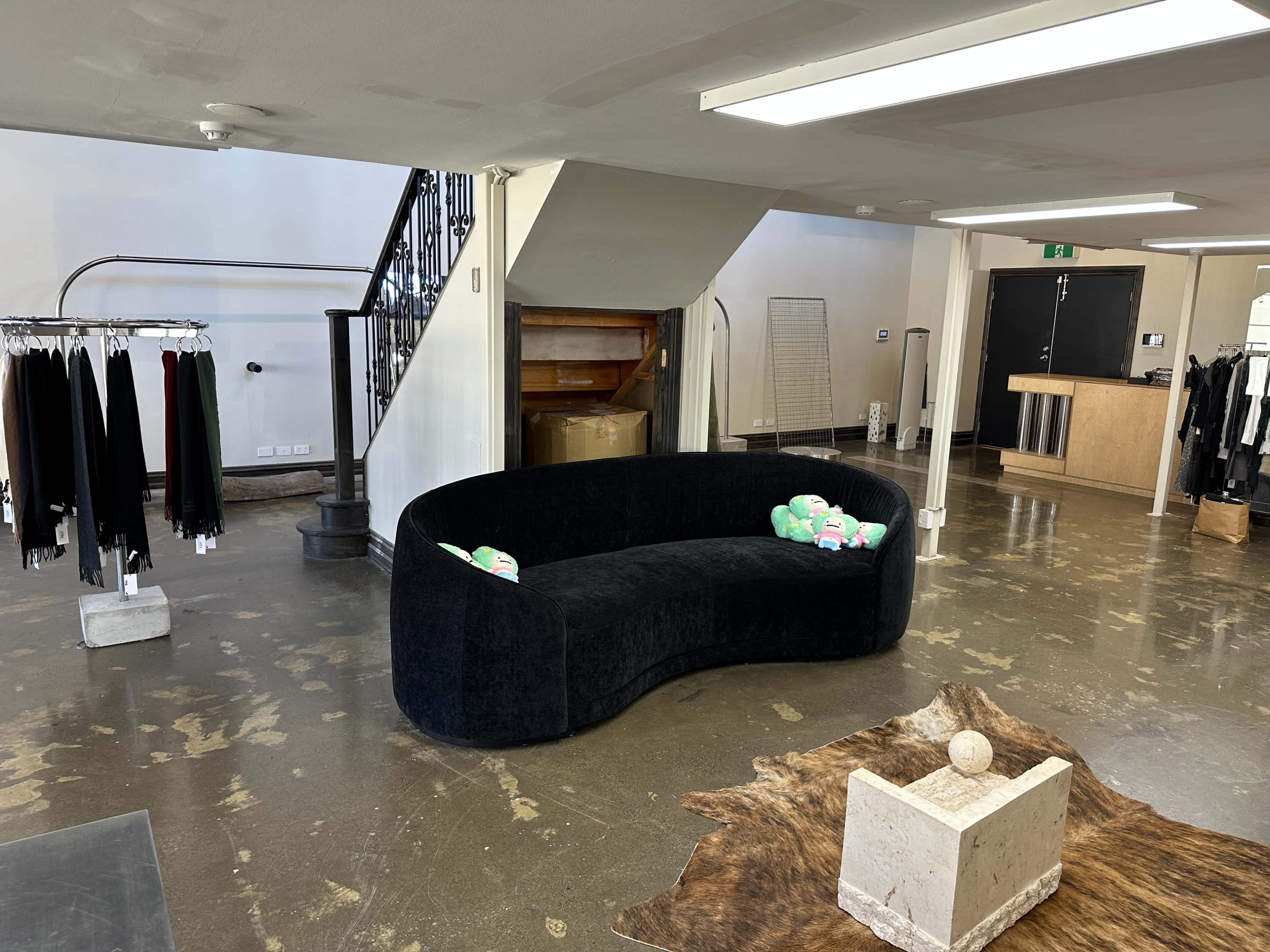 The image shows a modern clothing store interior featuring a curved black couch, a staircase in the background, and clothing racks to the left.