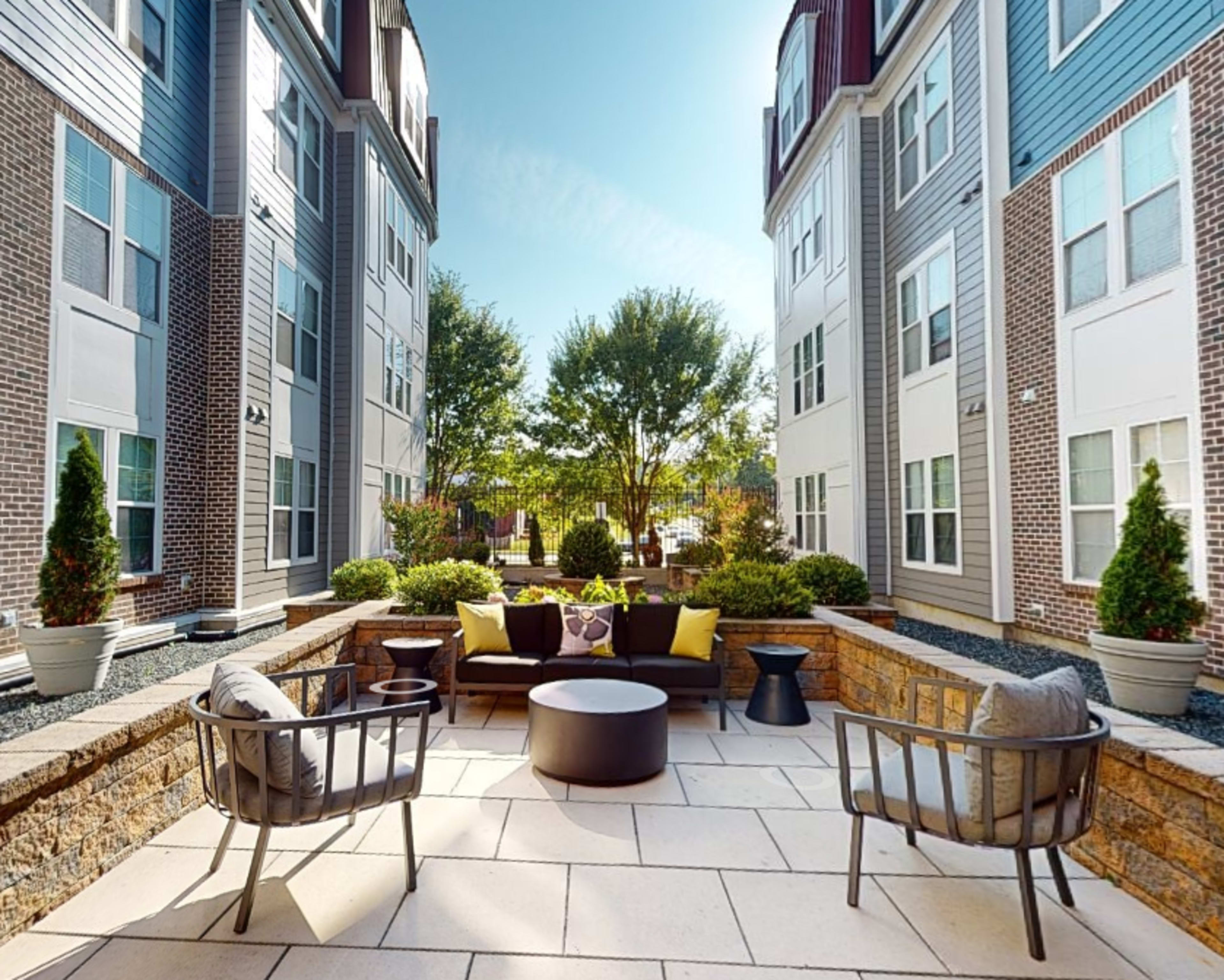 A landscaped outdoor seating area is enclosed by two multi-story apartment buildings, featuring circular and rectangular tables surrounded by cushioned chairs.