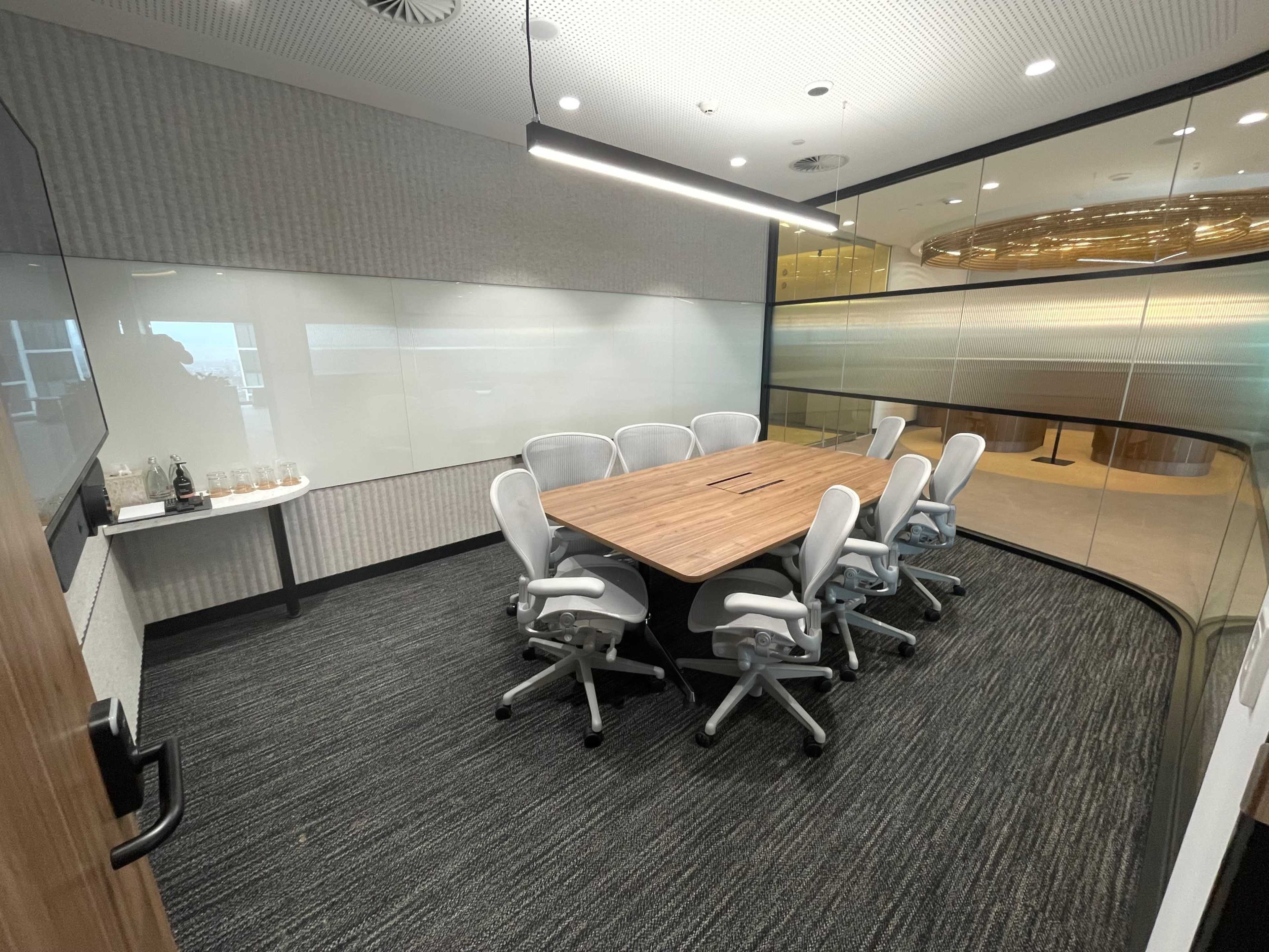 A conference room features a wooden table surrounded by six ergonomic chairs, with glass walls and a modern design.