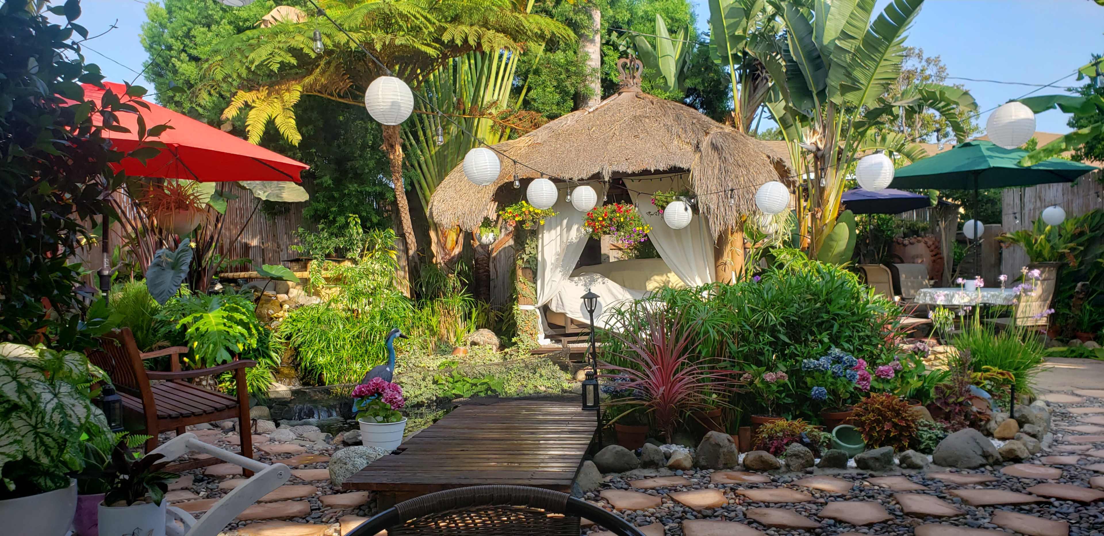 A landscaped garden features a thatched-roof hut surrounded by various plants, flowers, and decorative lanterns, with multiple seating areas and umbrellas.