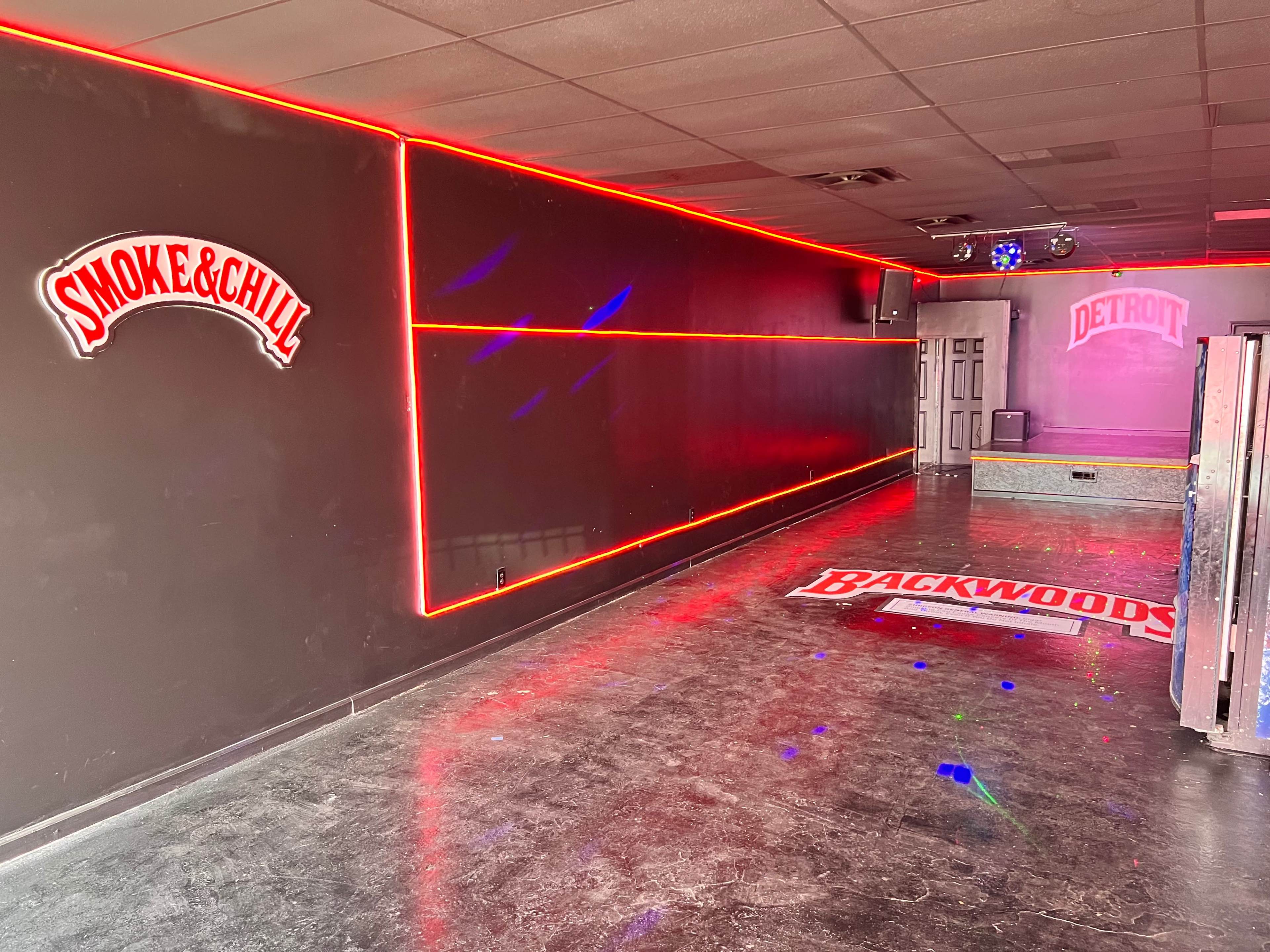 An empty interior space with dim lighting, neon accents along the walls, and signs for "Smoke & Chill" and "Backwoods."