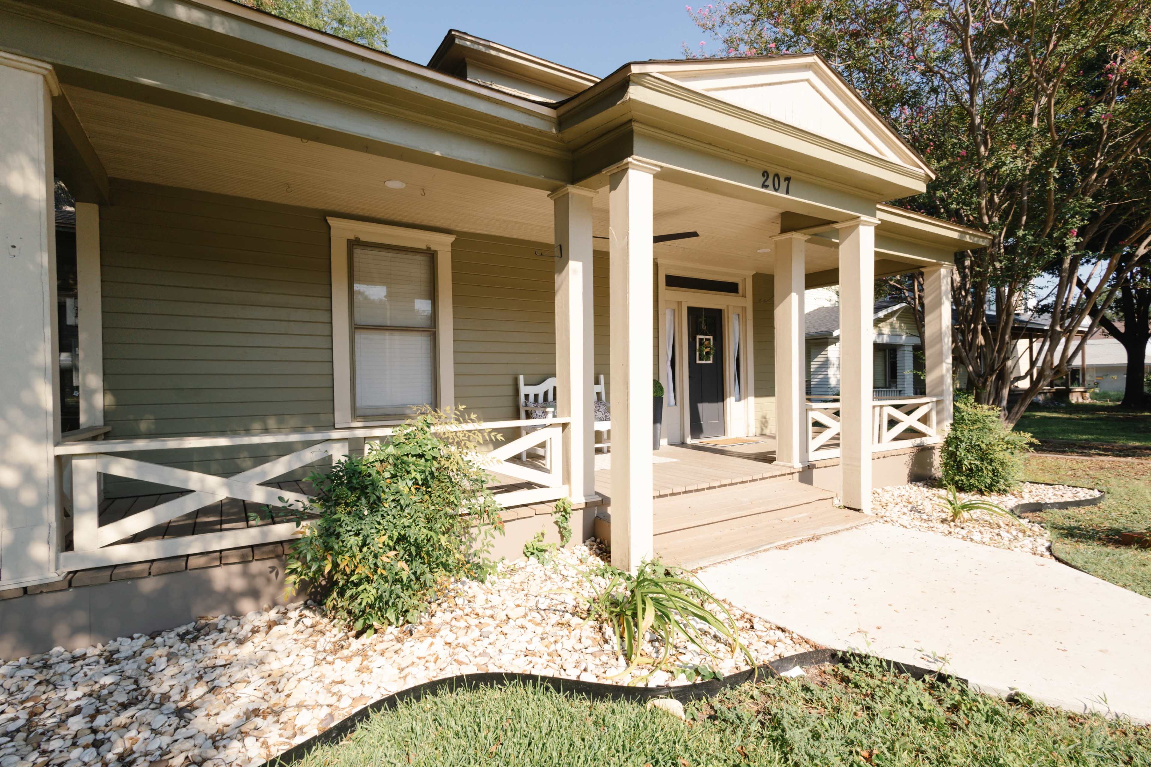 The image shows a single-story house with a covered porch, white rocking chairs, and a landscaped front yard with stones and greenery.
