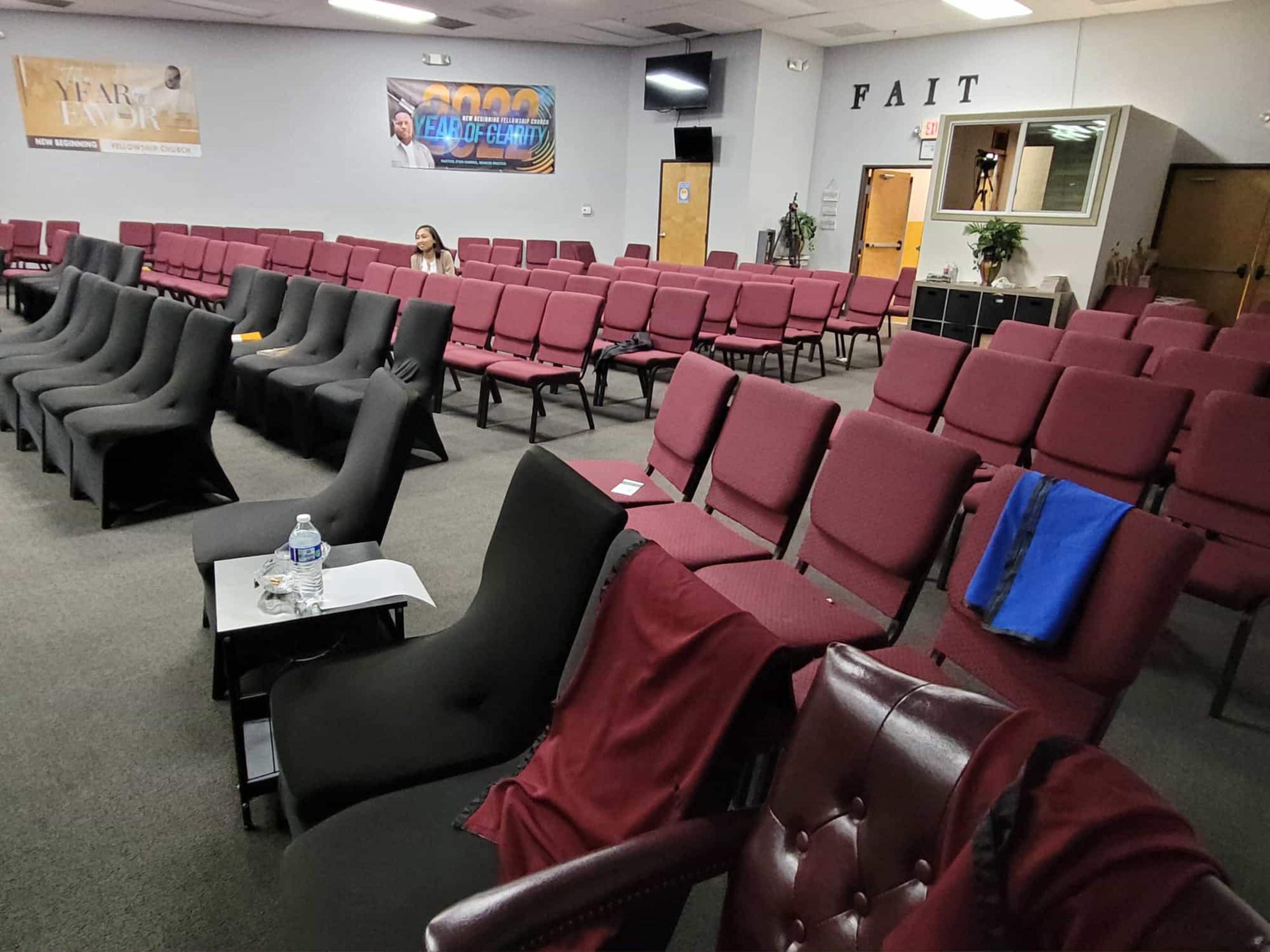 The image shows a nearly empty auditorium with rows of maroon chairs arranged in a formal layout, including a table with a water bottle in the foreground.