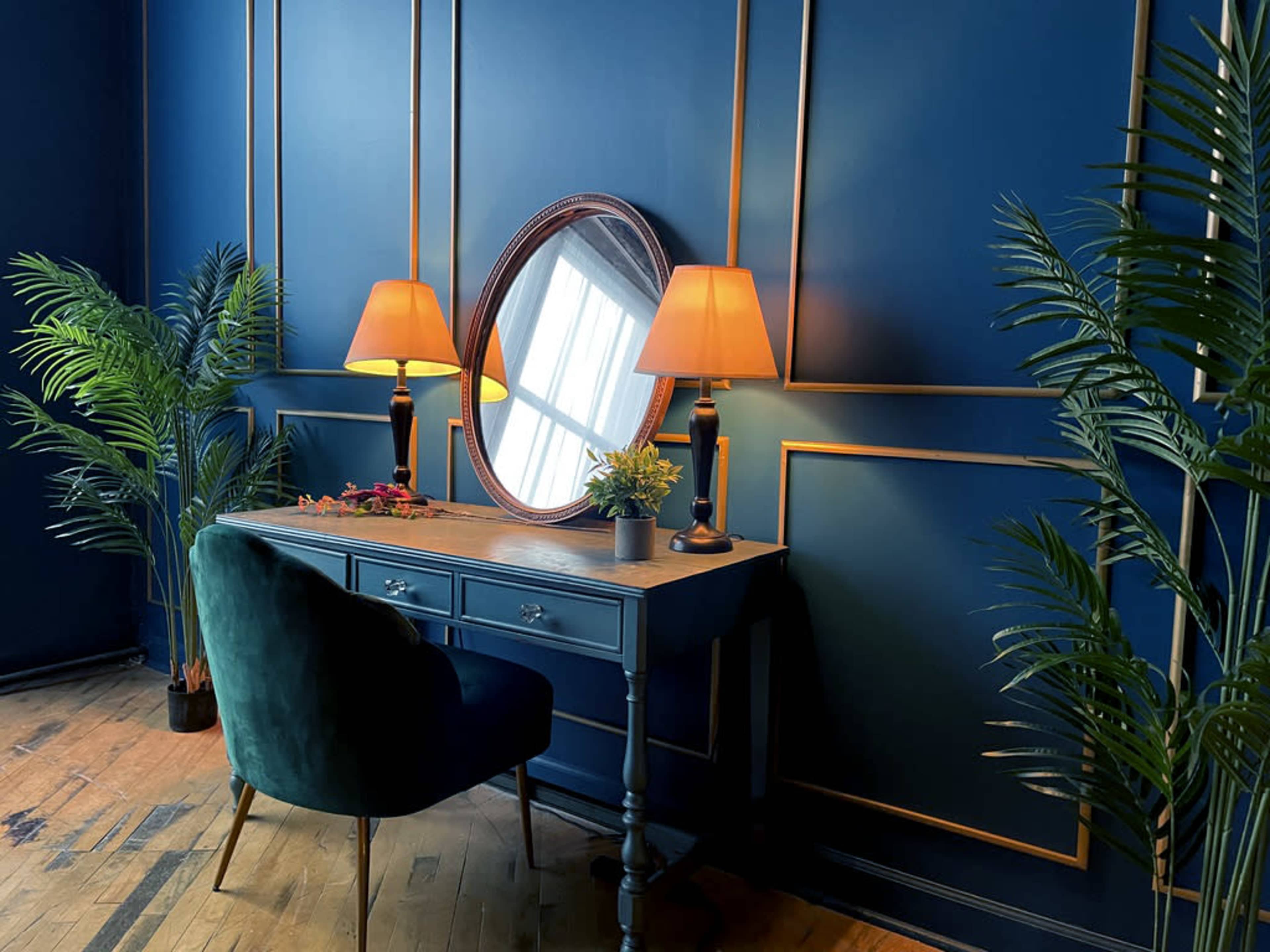 A blue-walled room features a dark wooden desk with a round mirror and two lamps beside potted plants.